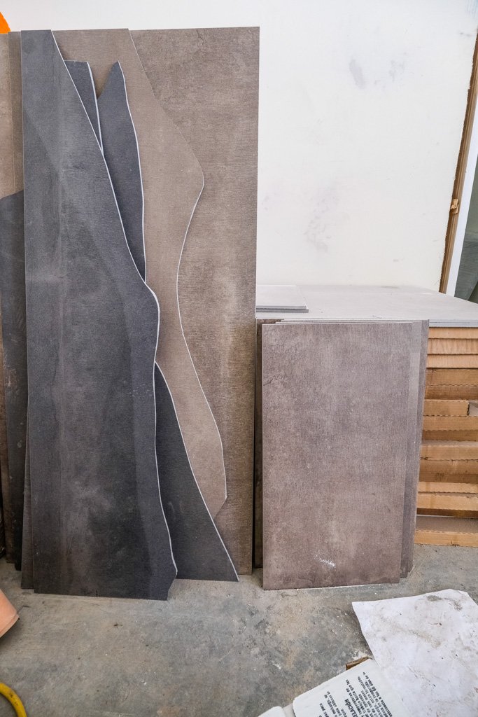 Stacked large rectangular tiles in shades of gray and beige leaning against a wall, with some stacked on a rack to the right. A paper and a yellow object are partially visible in the foreground.