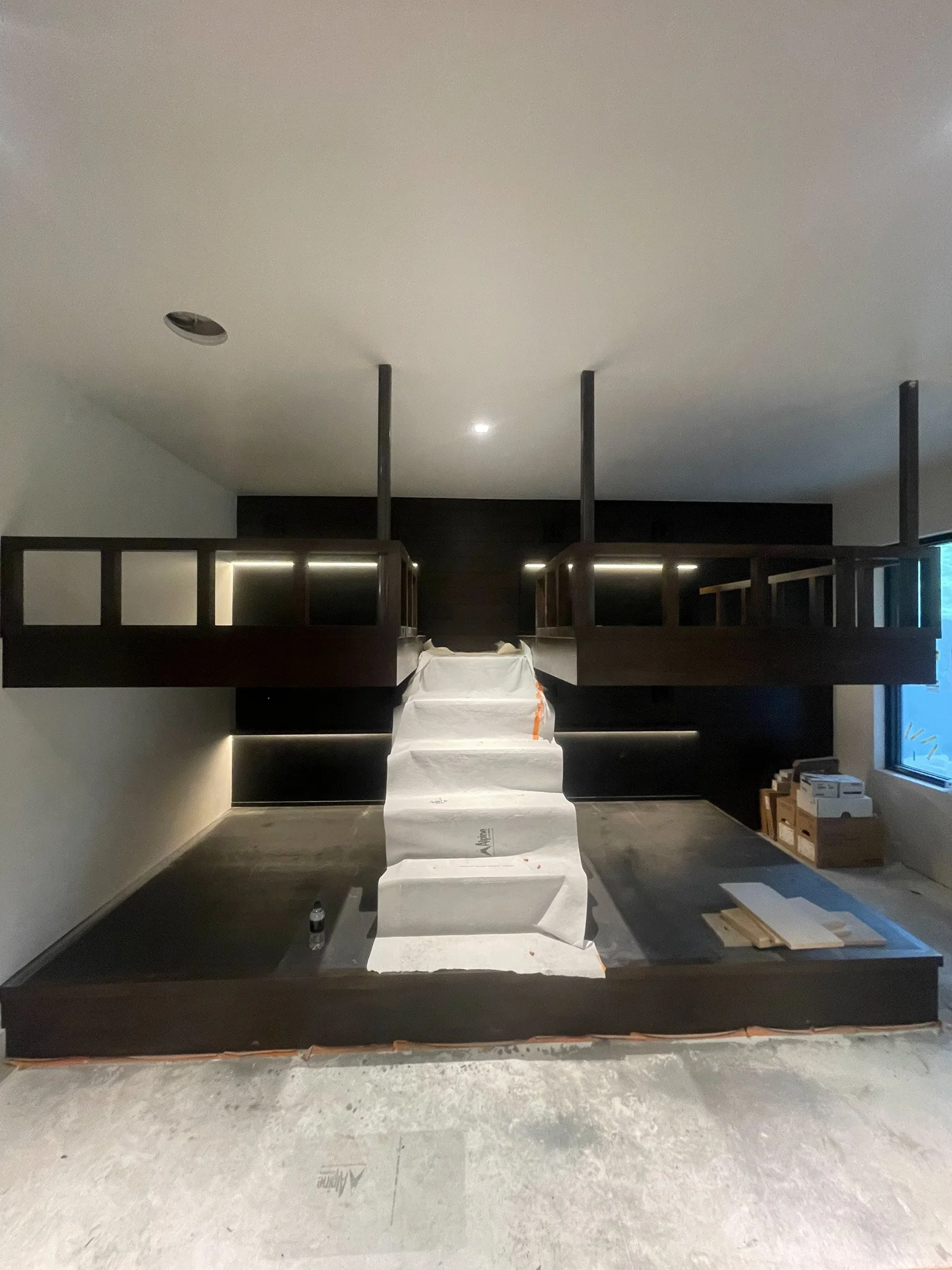 Interior of a room under construction featuring a raised platform with a staircase in the middle, surrounded by black walls and a large window on the right side.