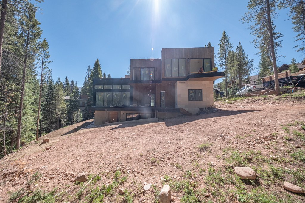 Modern multi-story house under construction on a dirt slope, surrounded by tall trees and other houses, with a clear blue sky overhead.
