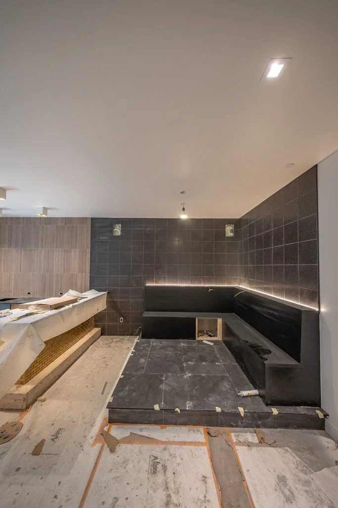 Interior space under construction, featuring black tiled seating area with LED lighting behind it and dark tiled walls, some construction materials and tools visible on the floor.