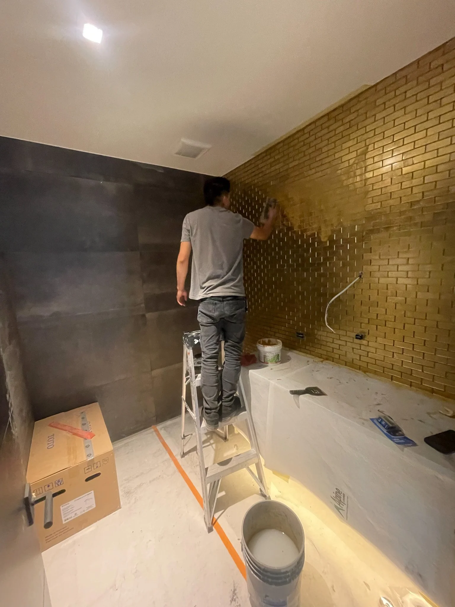 A person standing on a step ladder painting a brick wall gold in a room under renovation. There are construction materials and tools scattered around, including a bucket of paint and cardboard boxes.