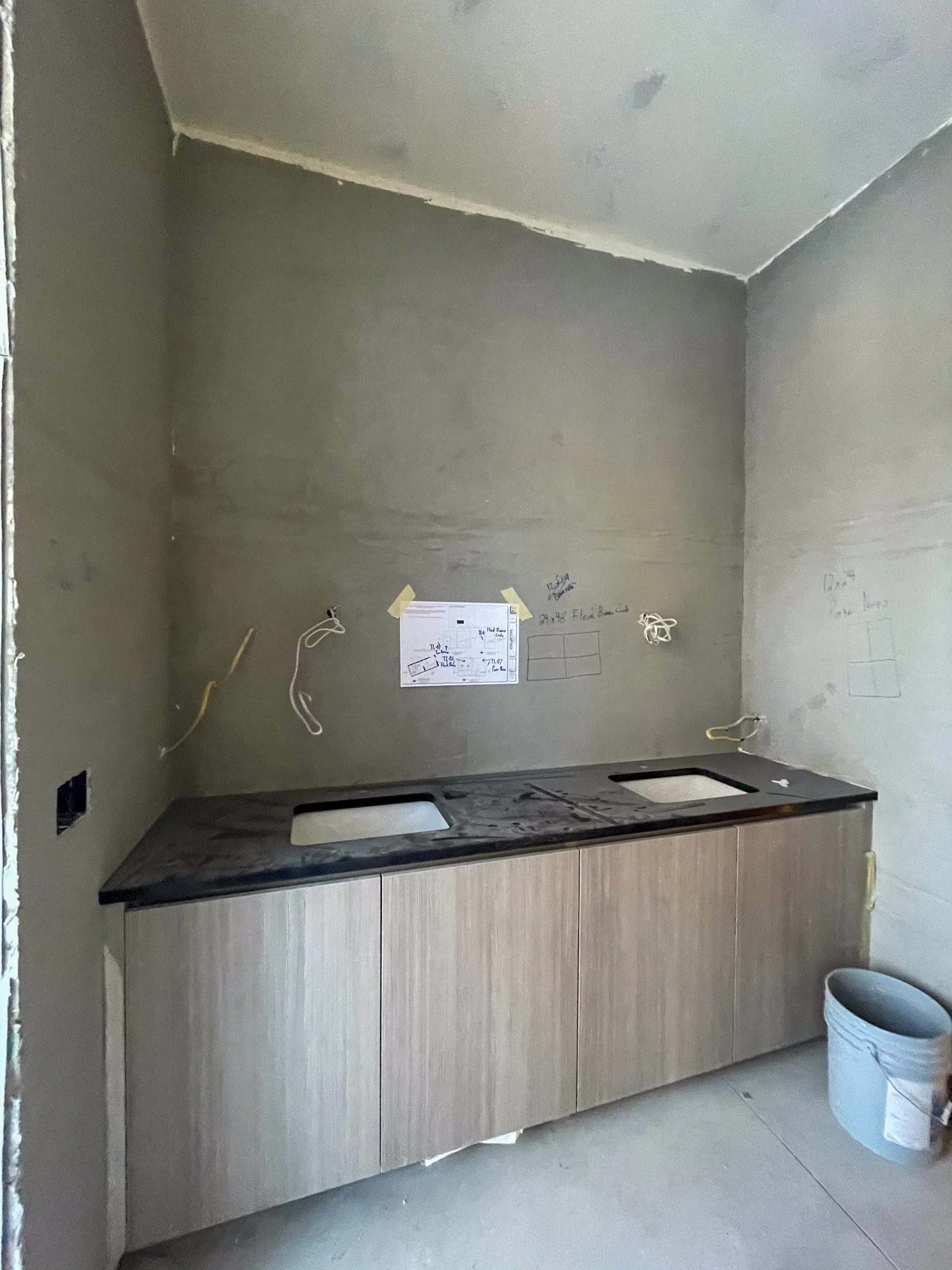 Incomplete kitchen with two sink bowls and a black countertop, unfinished walls with wiring and sketches, a bucket on the floor.