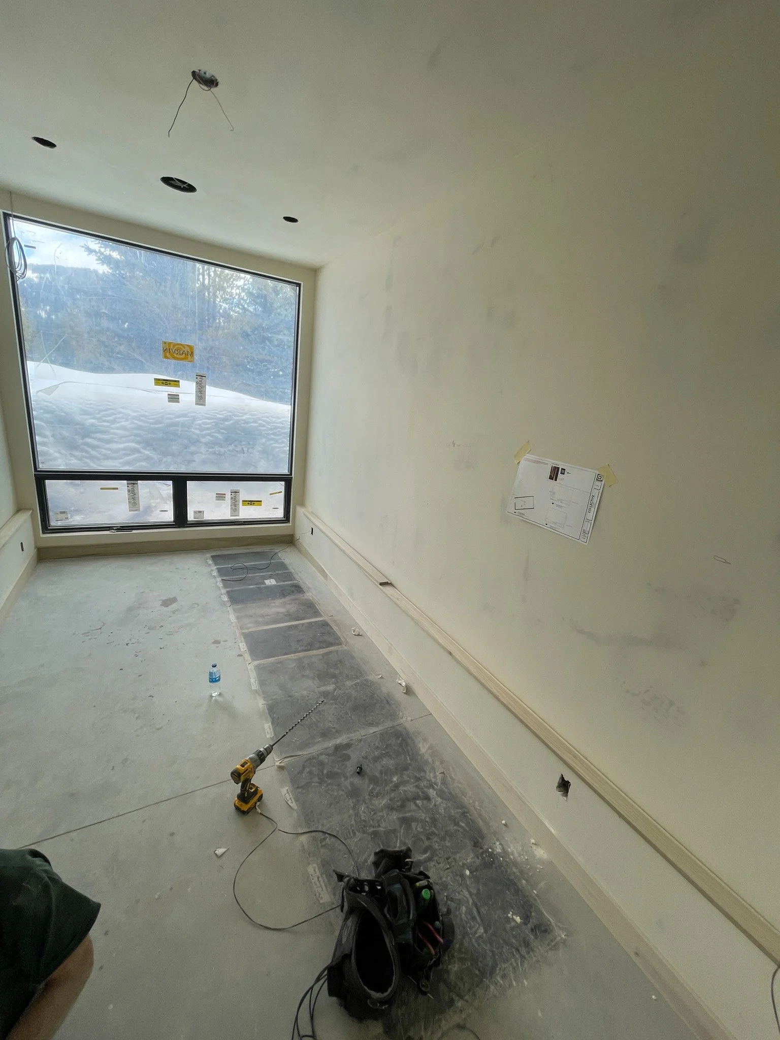 Interior of a room under construction or renovation, with unfinished walls, a large window showing a snowy landscape outside, construction tools on the floor, and electrical wiring hanging from the ceiling.