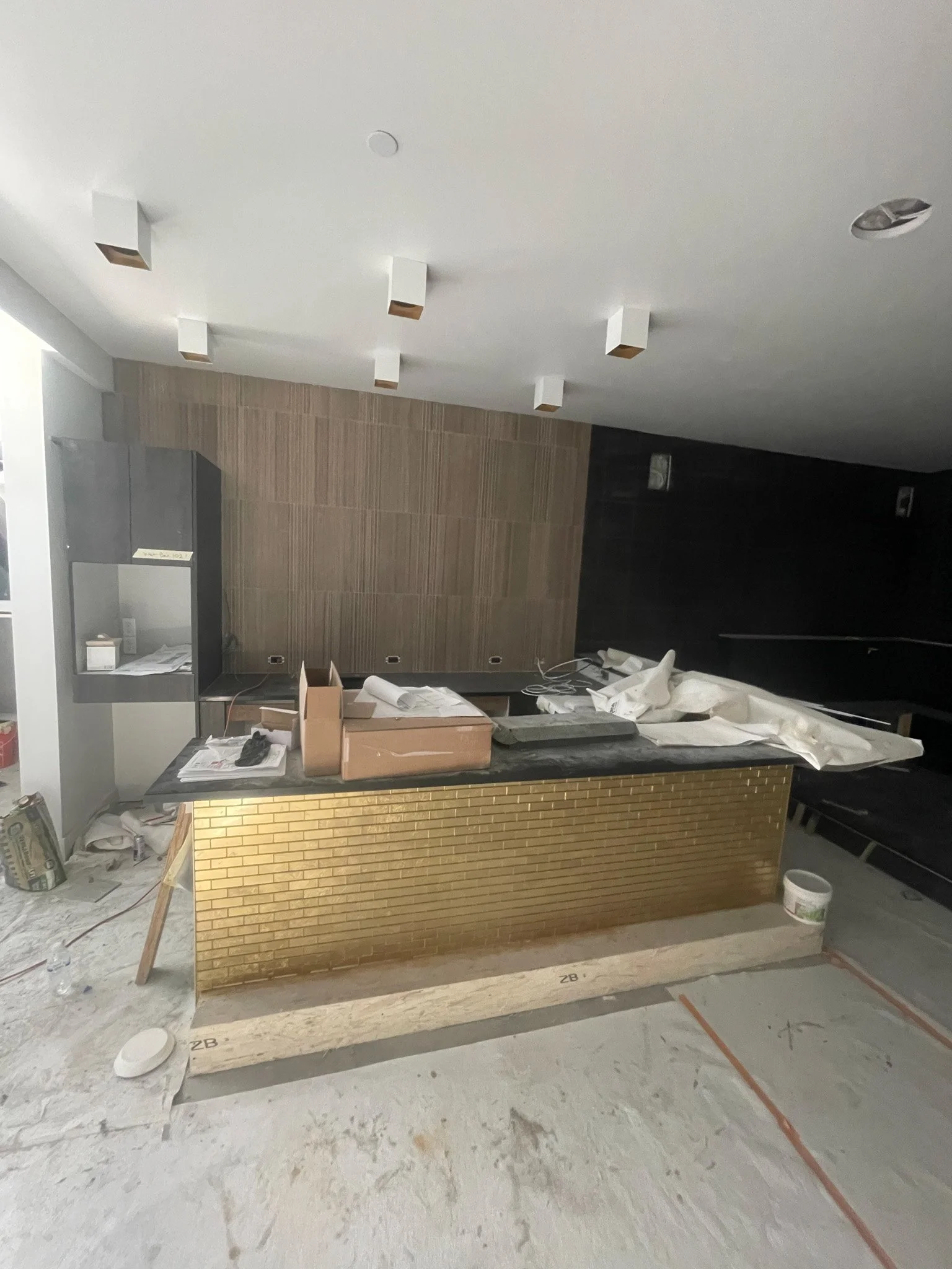 Interior of a construction or renovation site with a kitchen island covered with tools, papers, and materials, and a partially finished room with debris on the floor.