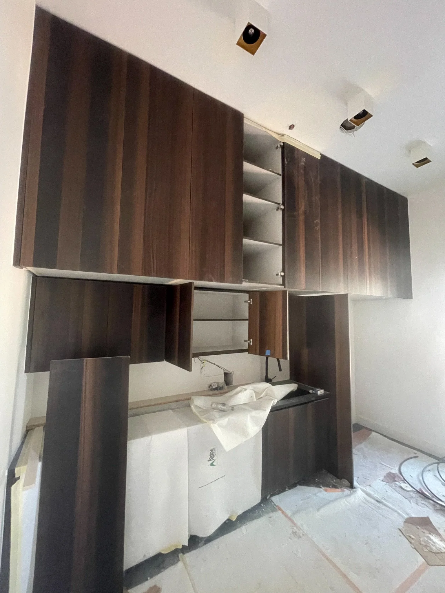 Kitchen cabinets under installation with dark wood finish, some open shelves and a counter space, surrounding a new refrigerator covered with protective film, in a room under construction.