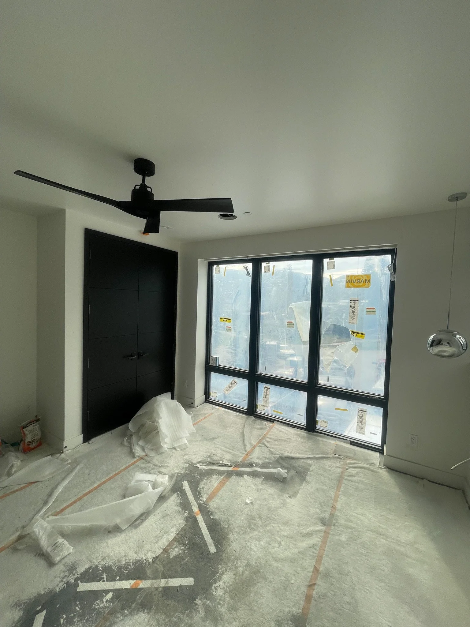 Empty room under construction with a black ceiling fan, large window, black closet doors, and construction materials on covered floor.