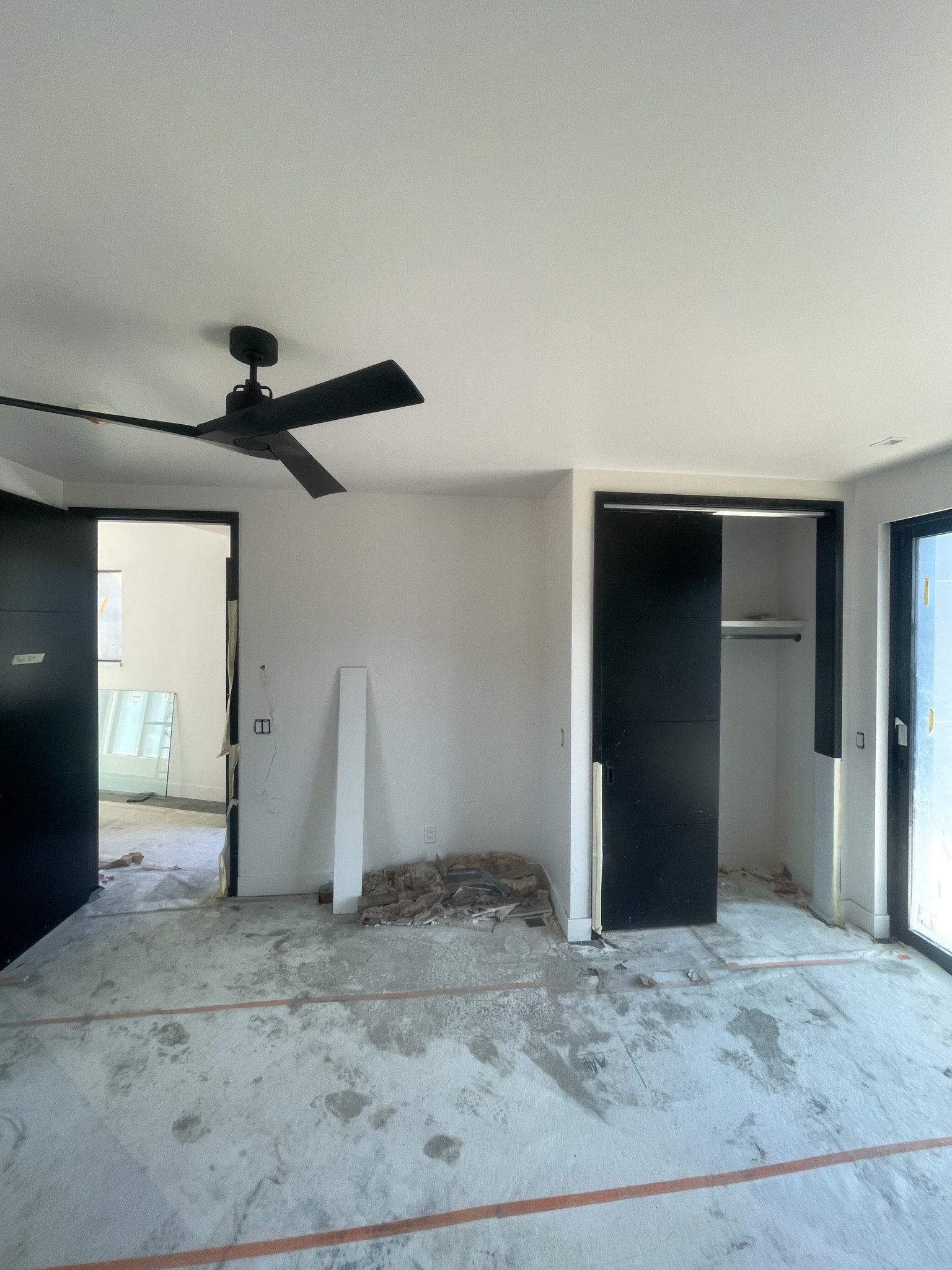 Room under construction with black closet and sliding door, unfinished flooring, and a black ceiling fan, leading into another room with windows.