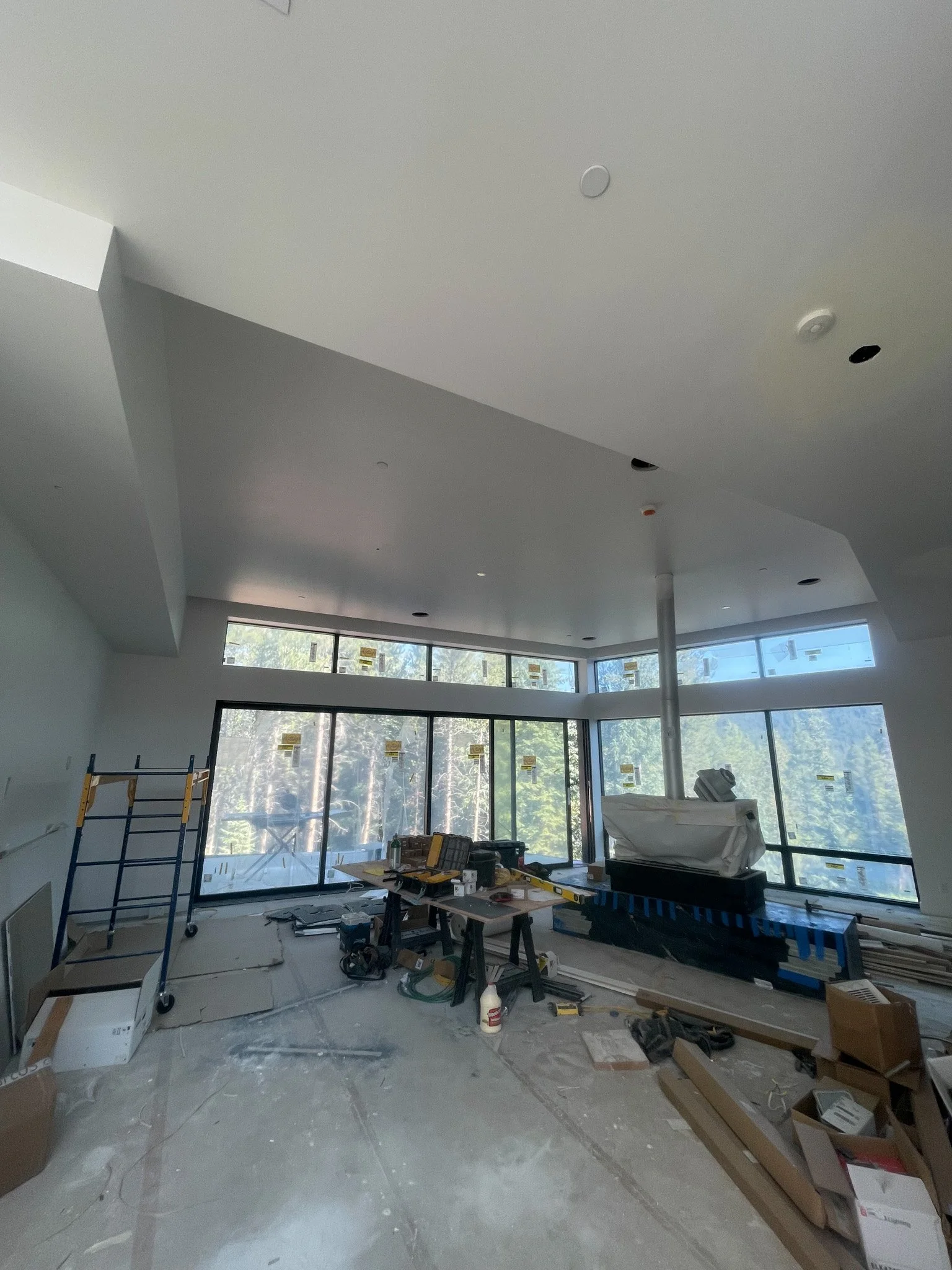Interior of a room under construction with large windows, construction tools, and materials scattered around, including a scaffold and covered equipment.