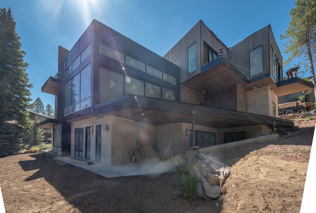 A modern, multi-story house under construction with large glass windows and wooden accents, situated on a sloped lot surrounded by trees and dirt, with construction workers visible on the upper levels.