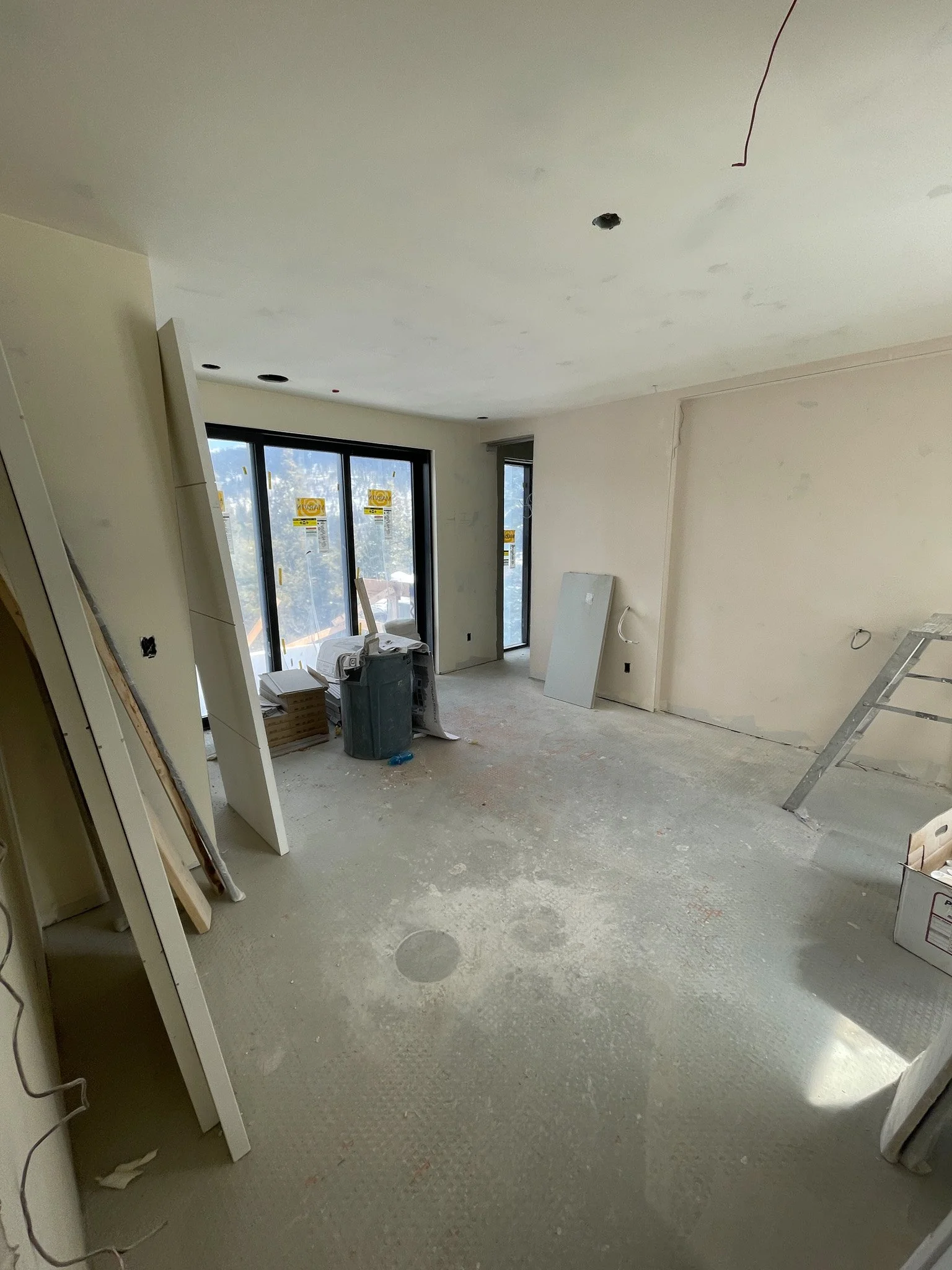 Interior of a room under construction with unfinished walls, ceiling, and floor. Construction materials and tools such as a ladder, cardboard boxes, and a trash bin are present. Large sliding glass doors leading outside are visible.