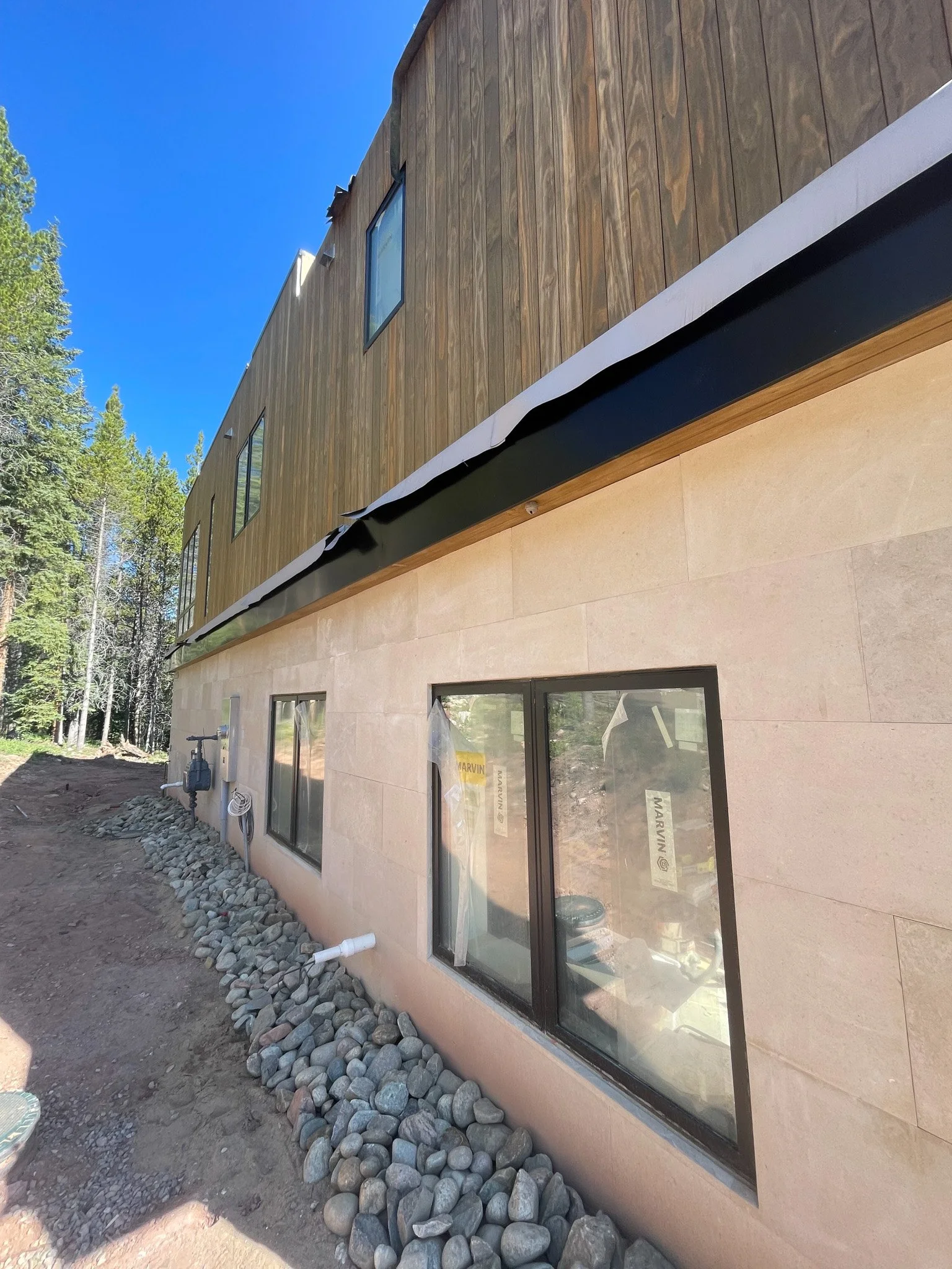 Exterior of a modern house under construction, showing a foundation with large rocks, windows, and a wooden upper level with some windows, set in a wooded area under a clear blue sky.