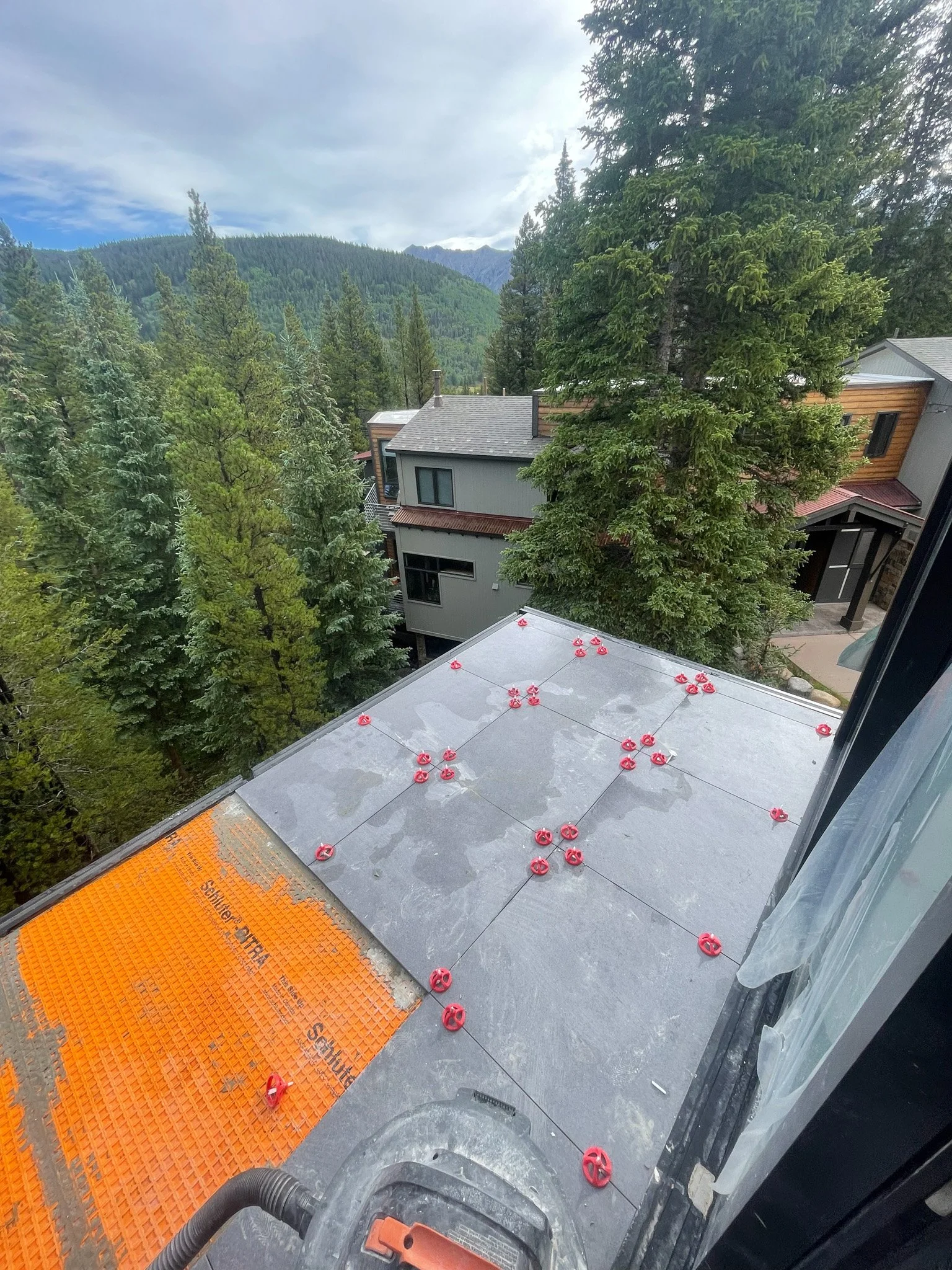 View from a construction or renovation site on a building's rooftop, showing grey roofing tiles being installed with red fasteners, surrounded by tall green trees and neighboring houses with mountain scenery in the background.