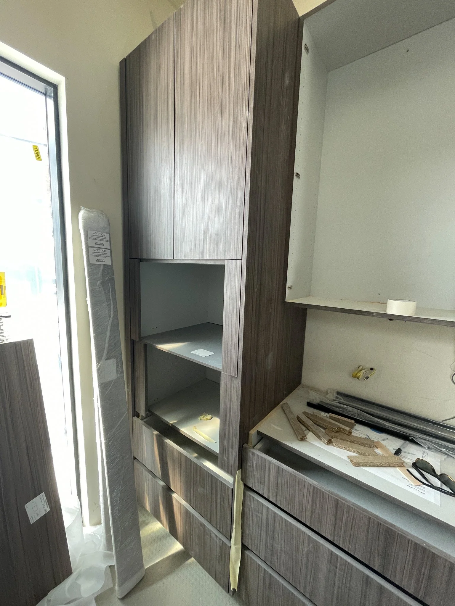 Kitchen cabinetry under construction with unfinished drawers and shelves, construction tools, and materials on the countertops, in a room with a door to the exterior on the left.