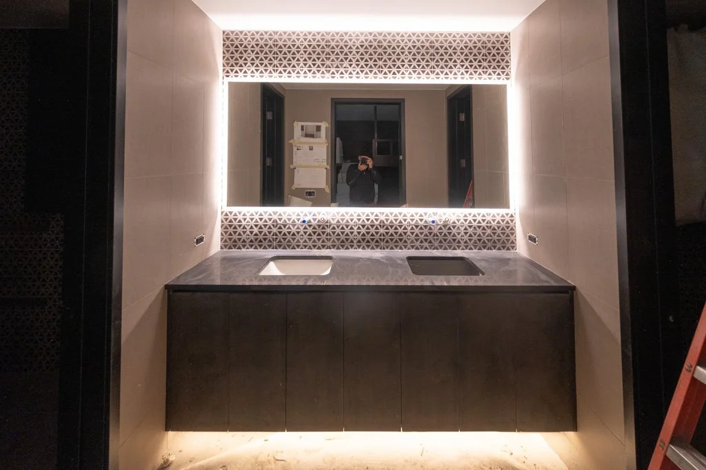 Modern bathroom with illuminated mirror, black vanity with dual sinks, beige tiled walls, and decorative triangle-patterned accent above the mirror.