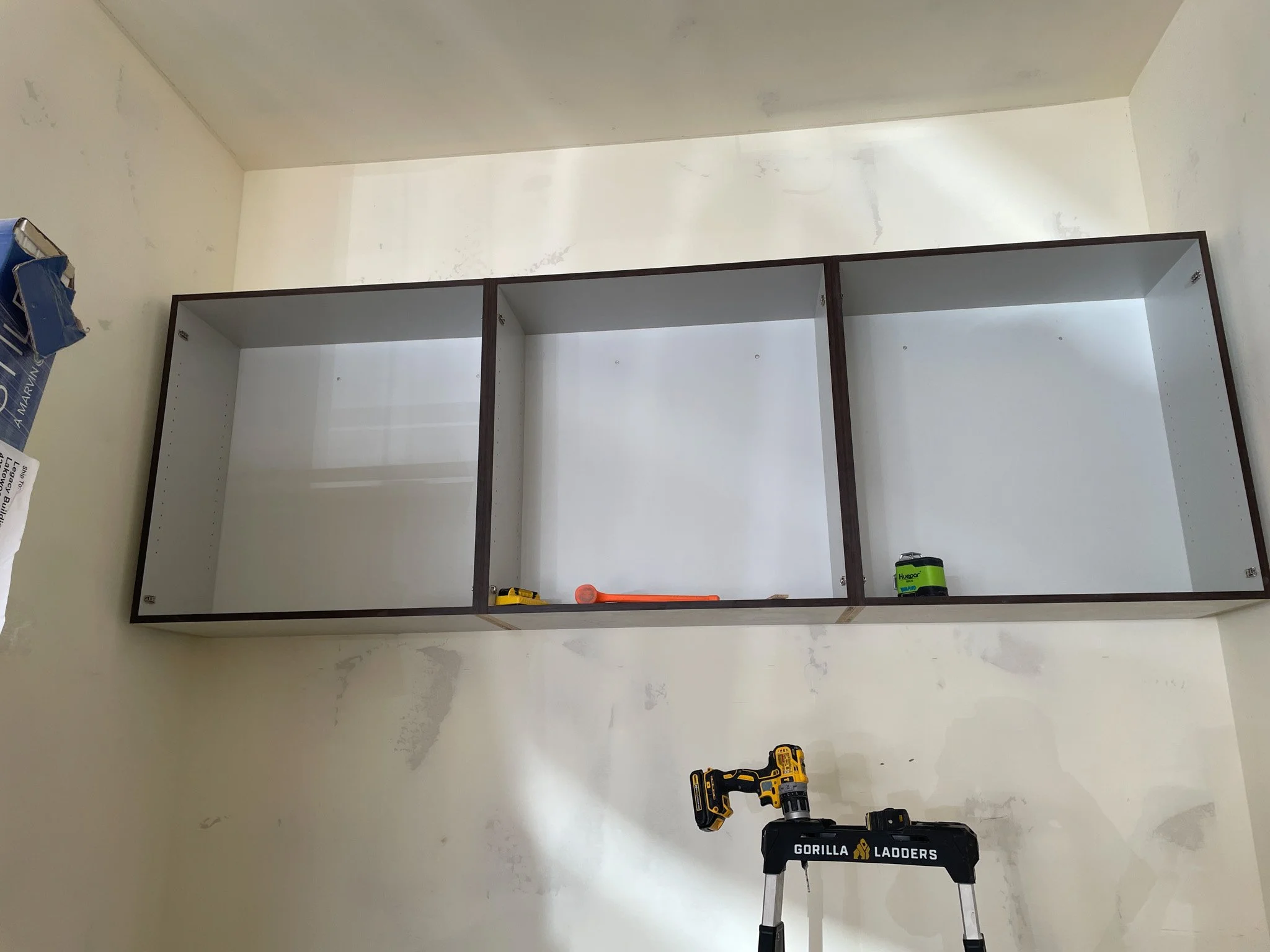 Empty three-compartment wall cabinet being installed on a white wall with tools and a drill stand nearby.