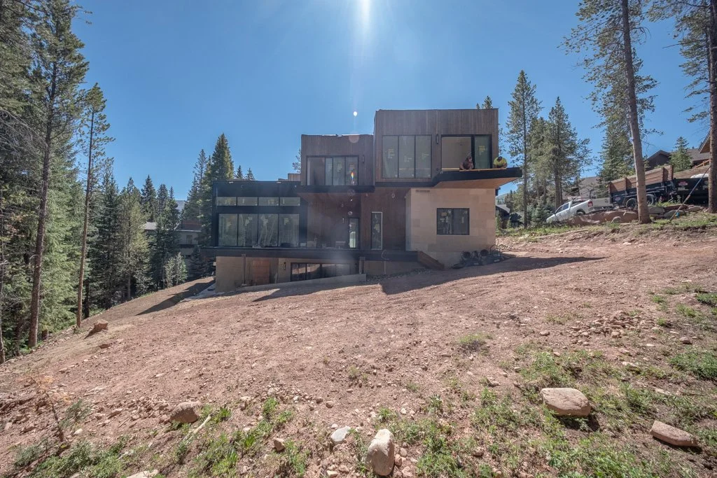 Modern house under construction on a sloped wooded lot, with trees and clear skies in the background.