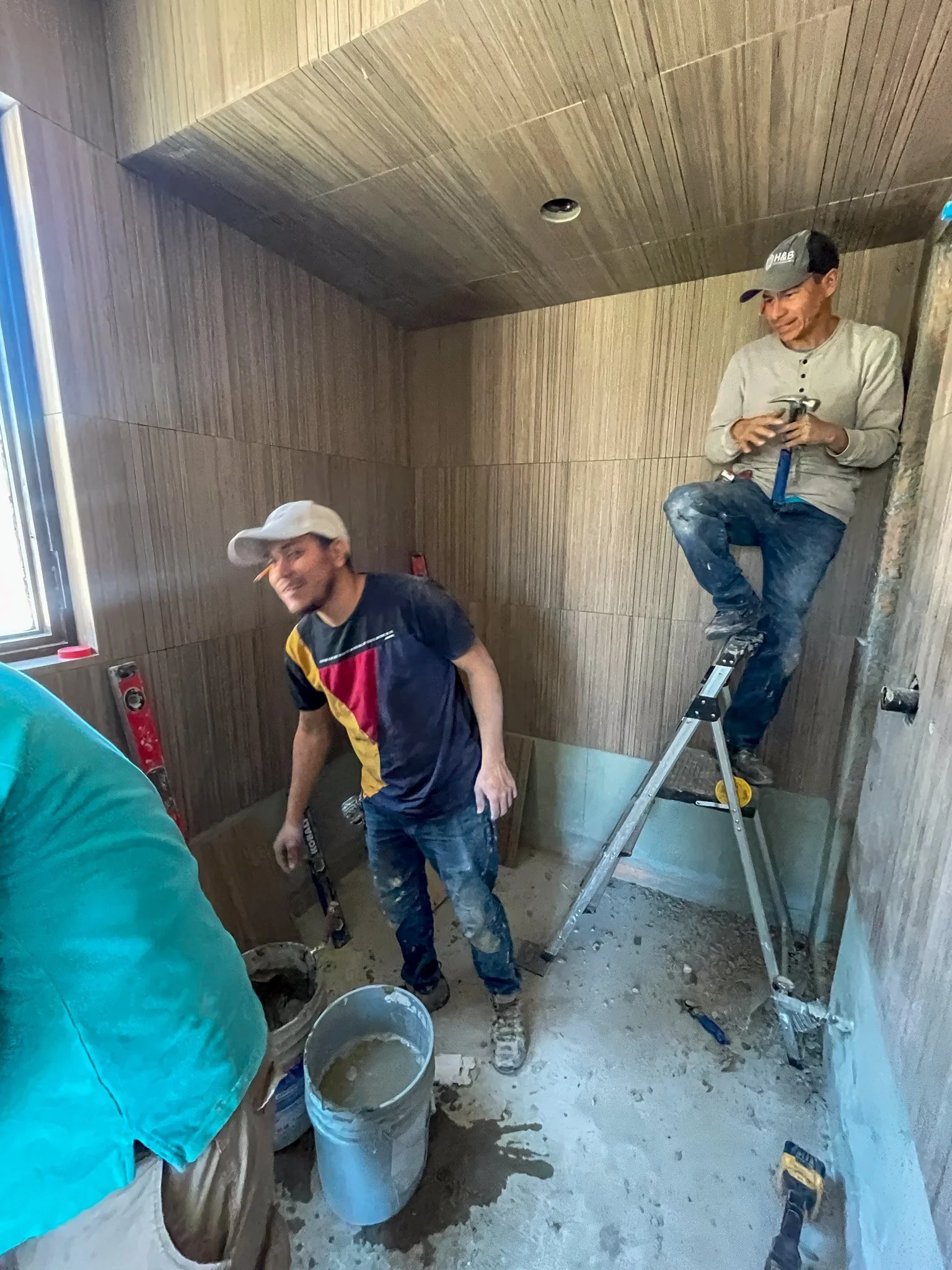 Two construction workers installing wall panels in a small room. One is standing on a ladder holding a tool, while the other is standing on the floor. The room is under renovation with construction materials and tools scattered around.