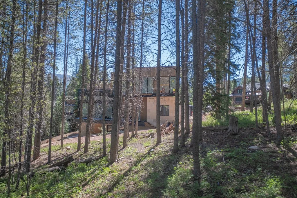 A house under construction in a wooded area, surrounded by trees and sunlight.