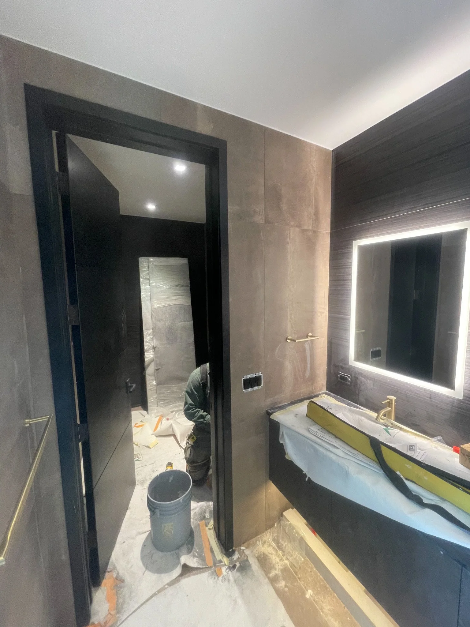 Bathroom under construction with a large illuminated mirror, a countertop with a sink, and workers installing fixtures.