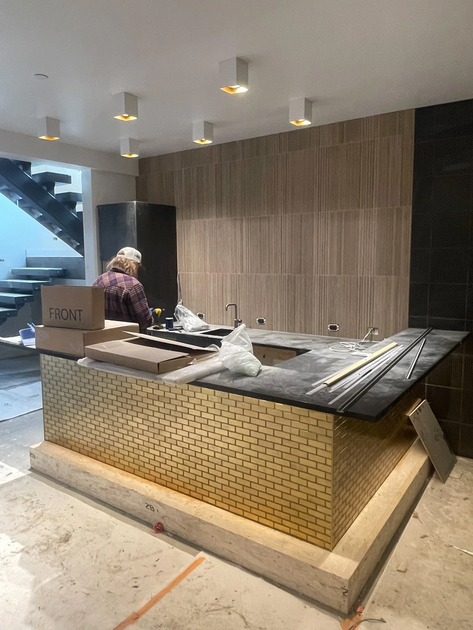 A kitchen or bar area under construction, featuring a brick-front island with a black countertop, partially assembled with construction materials and tools on top, and a worker in a plaid shirt and cap working in the background.