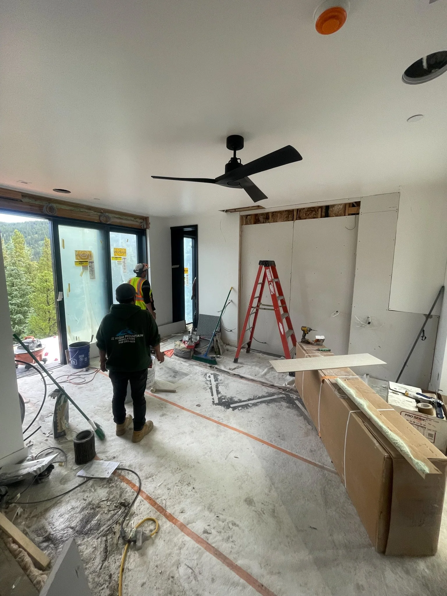 Interior view of a room under construction with two workers, construction tools, a ladder, and exposed wall area.