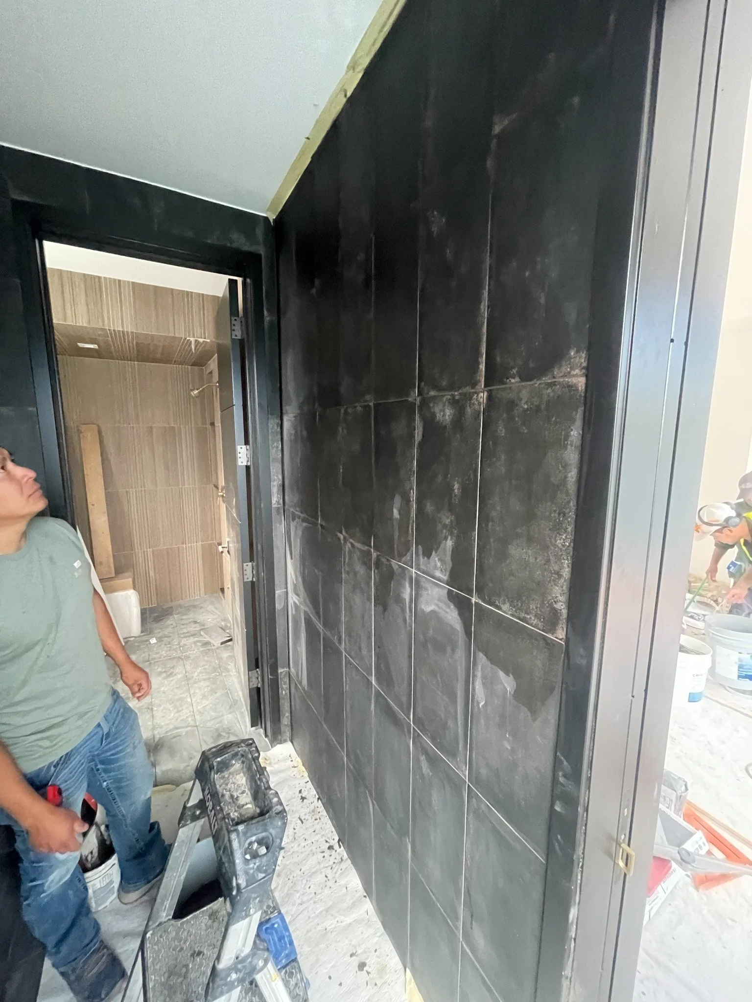 A construction worker standing next to a partially tiled black wall, with construction tools and materials around, in a room under renovation.