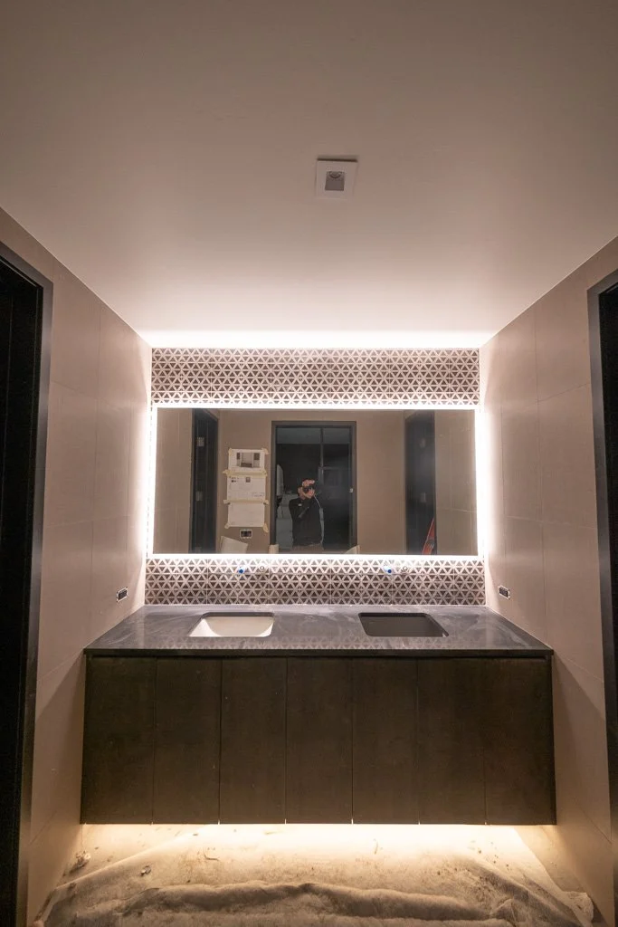 Modern bathroom with a large illuminated mirror above a black vanity with a sink and cooktop, beige tiled walls, and a decorative upper trim.
