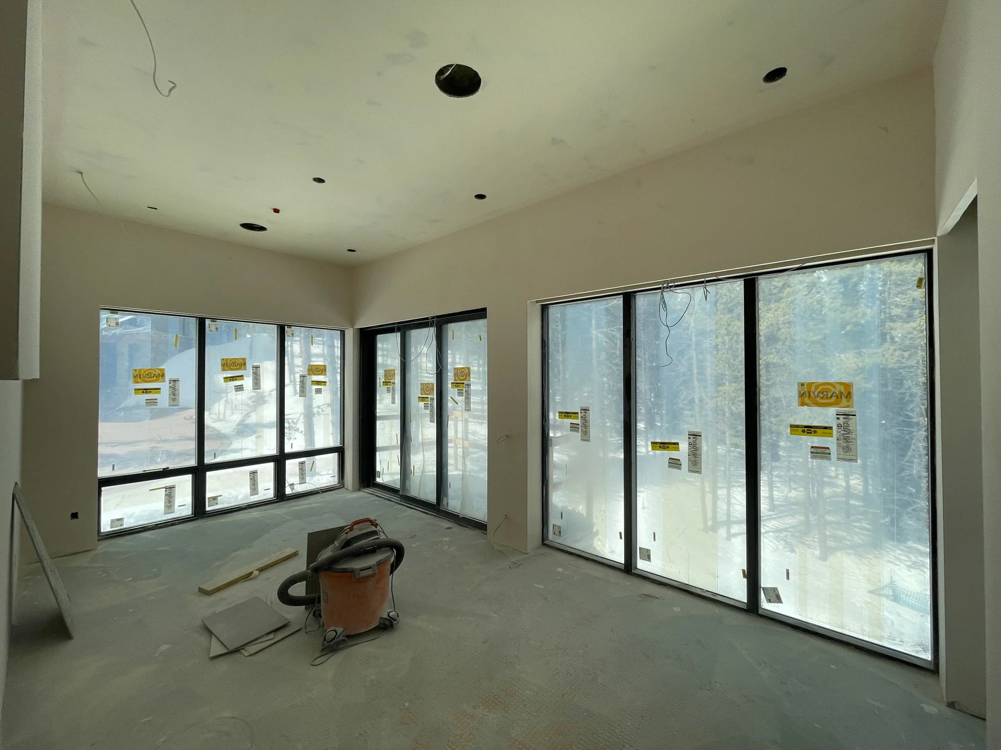 Room under construction with large windowed sliding glass doors, unfinished ceiling with holes, construction tools on floor, and snow visible outside.