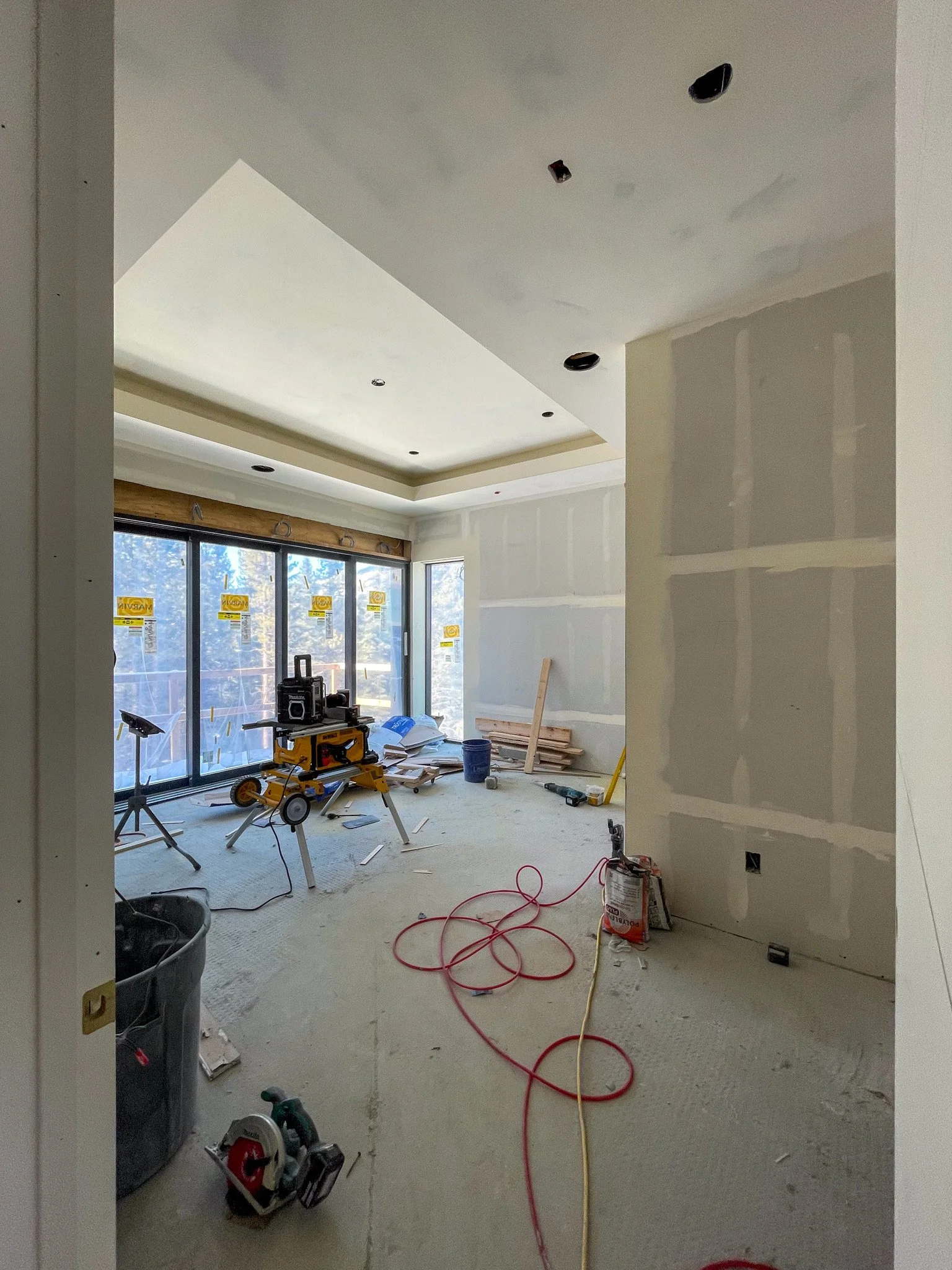 Interior view of a room under construction with tools, construction materials, and partially finished drywall, large window with stickers, and no ceiling fixtures installed.