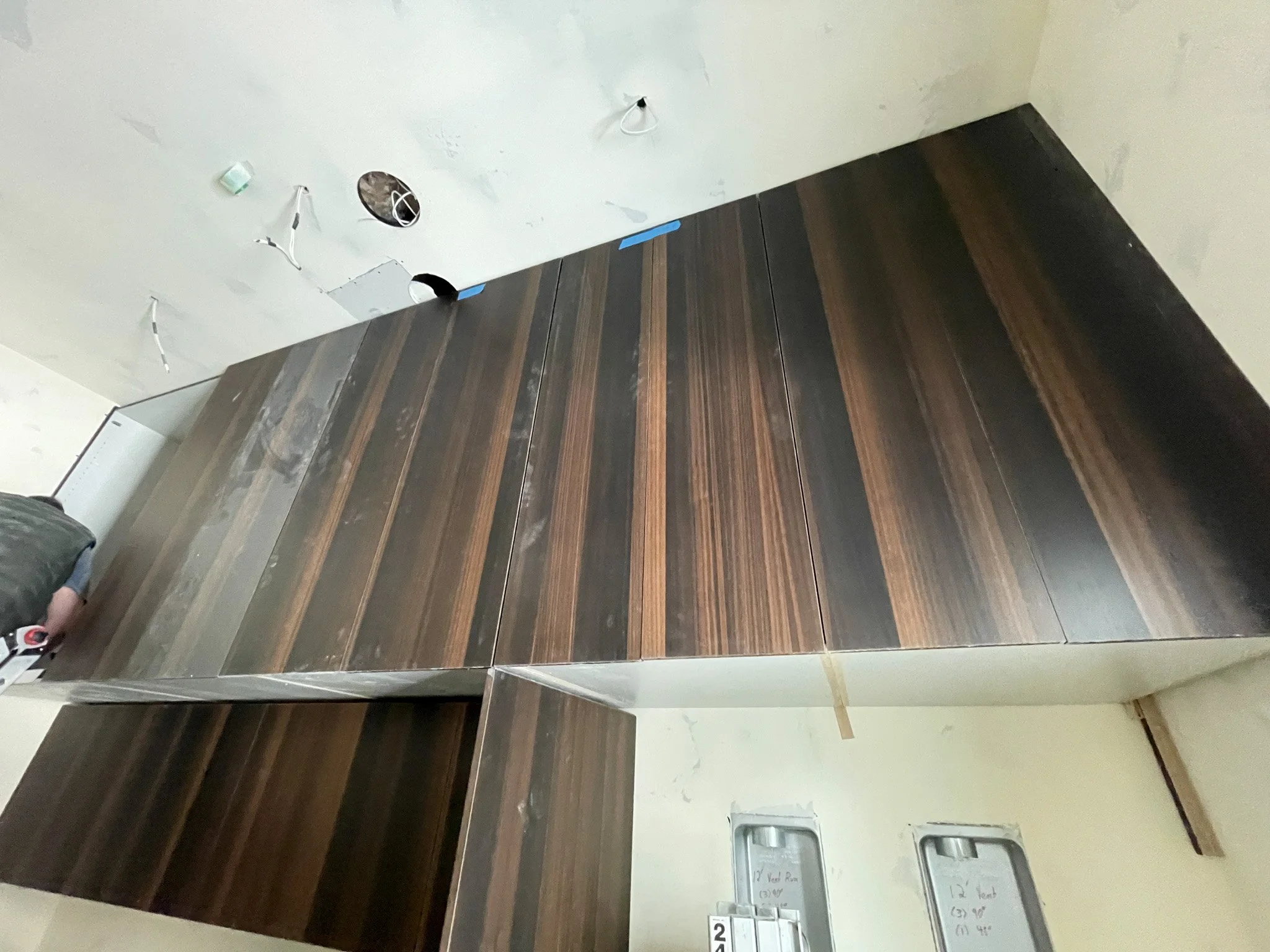 Interior of a room under construction, with dark wooden cabinets or panels. The ceiling has holes and wires hanging, with a few small fixtures installed.