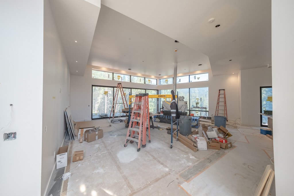 Interior space under construction with ladders, construction tools, and materials scattered around, large windows at the front, and white walls and ceiling.