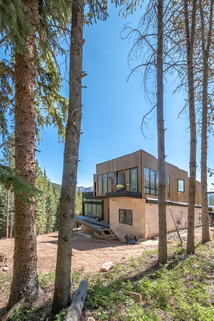 Contemporary house under construction in a wooded area, surrounded by tall pine trees, with exterior wood and stone finishes, large windows, and a construction worker visible on the upper level.