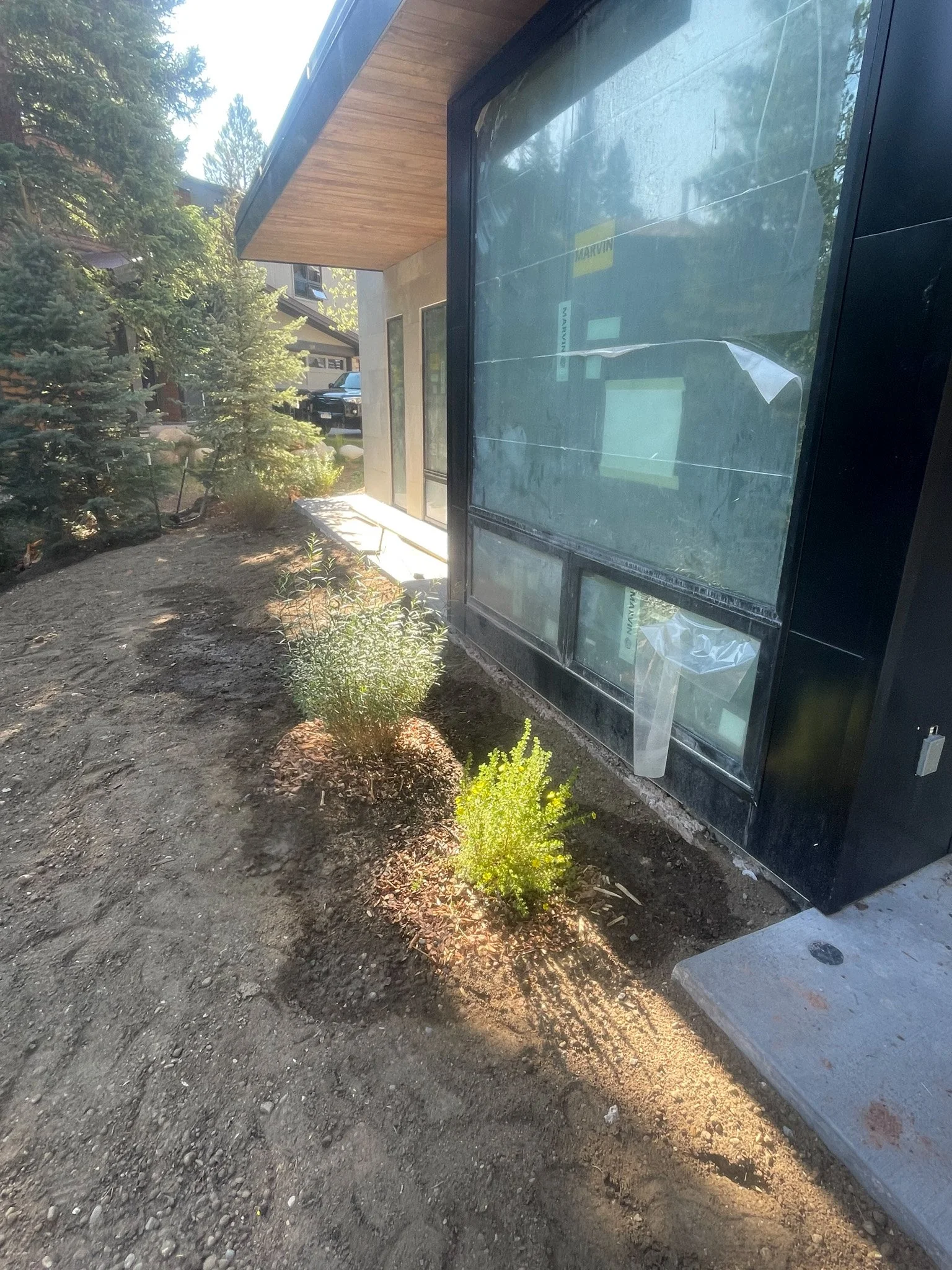 Exterior view of a house under construction, showing a large glass window, wooden roof overhang, and young shrubs planted in the soil beside the house.