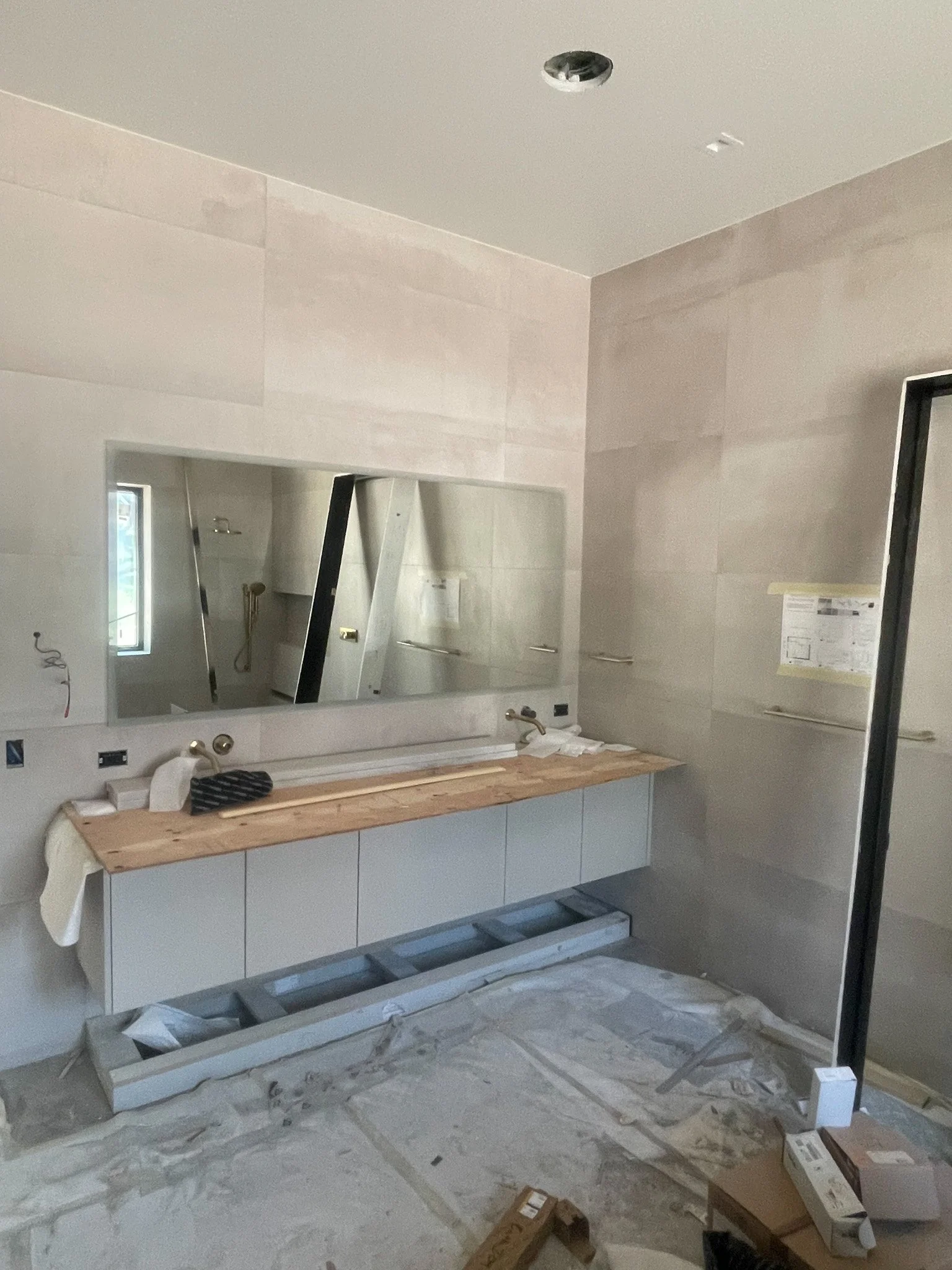 A bathroom under construction with a double vanity with drawer cabinet and a large mirror above. The countertop is wood, and there are fixtures for two sinks. The wall is tiled, and construction materials and tools are scattered on the floor.