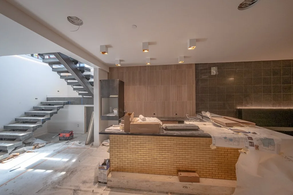 Interior of a house under construction with a staircase on the left, a kitchen island with yellow brick facade, and construction materials scattered throughout.