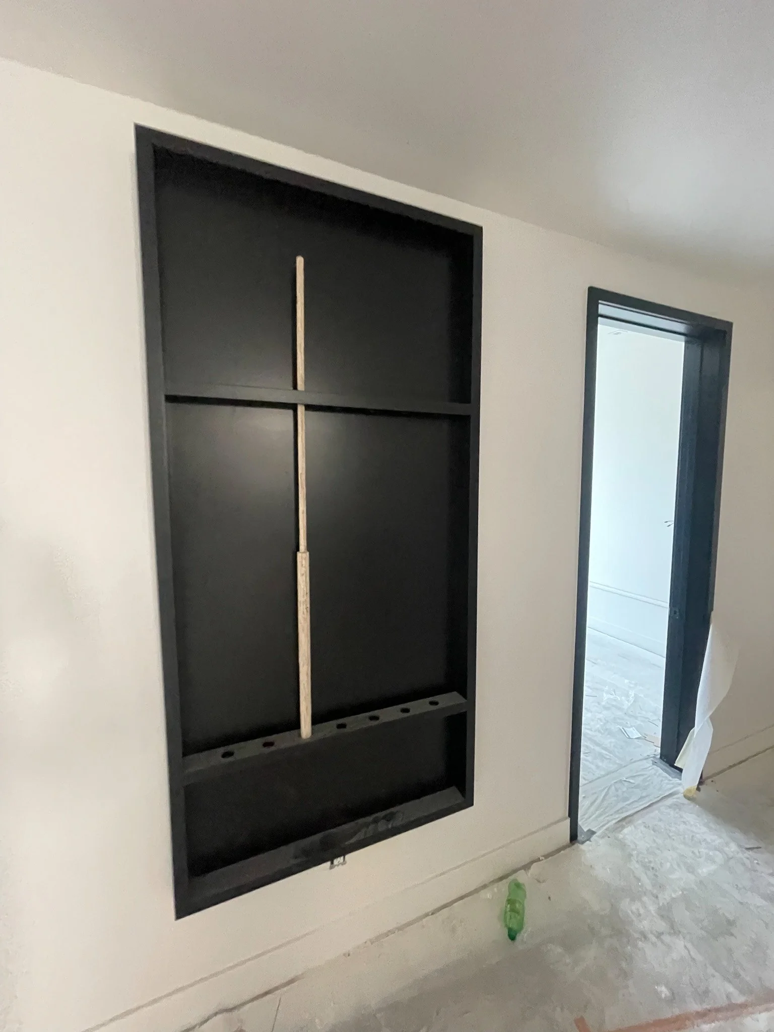 Interior wall with a black recessed shelving unit and an open doorway leading to a room with construction materials on the floor.