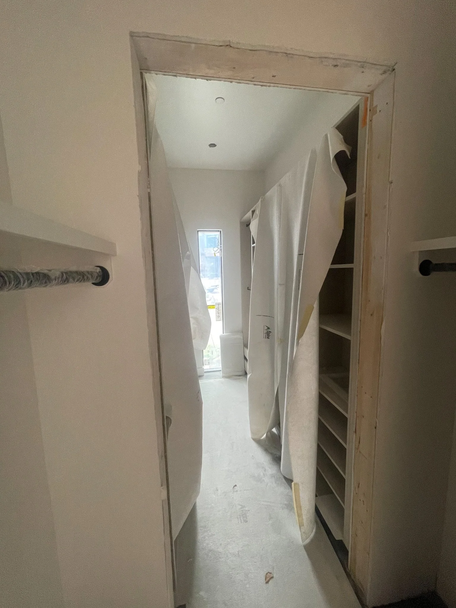 A walk-in closet under construction with partially installed shelves on the right side and hanging rods on the left side. The doorway frame is unfinished, showing exposed wood and drywall. In the background, through a door or window, a bright outdoor