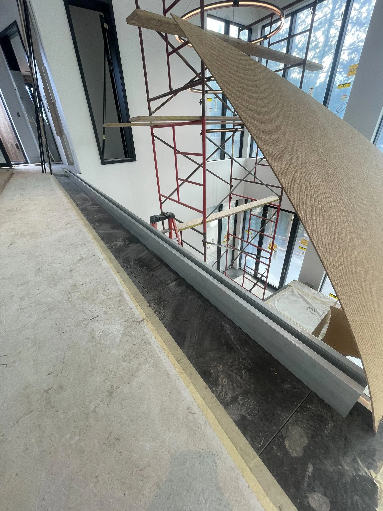 Interior of a building under construction with scaffolding, a construction barrier, and partially finished flooring near a large window.