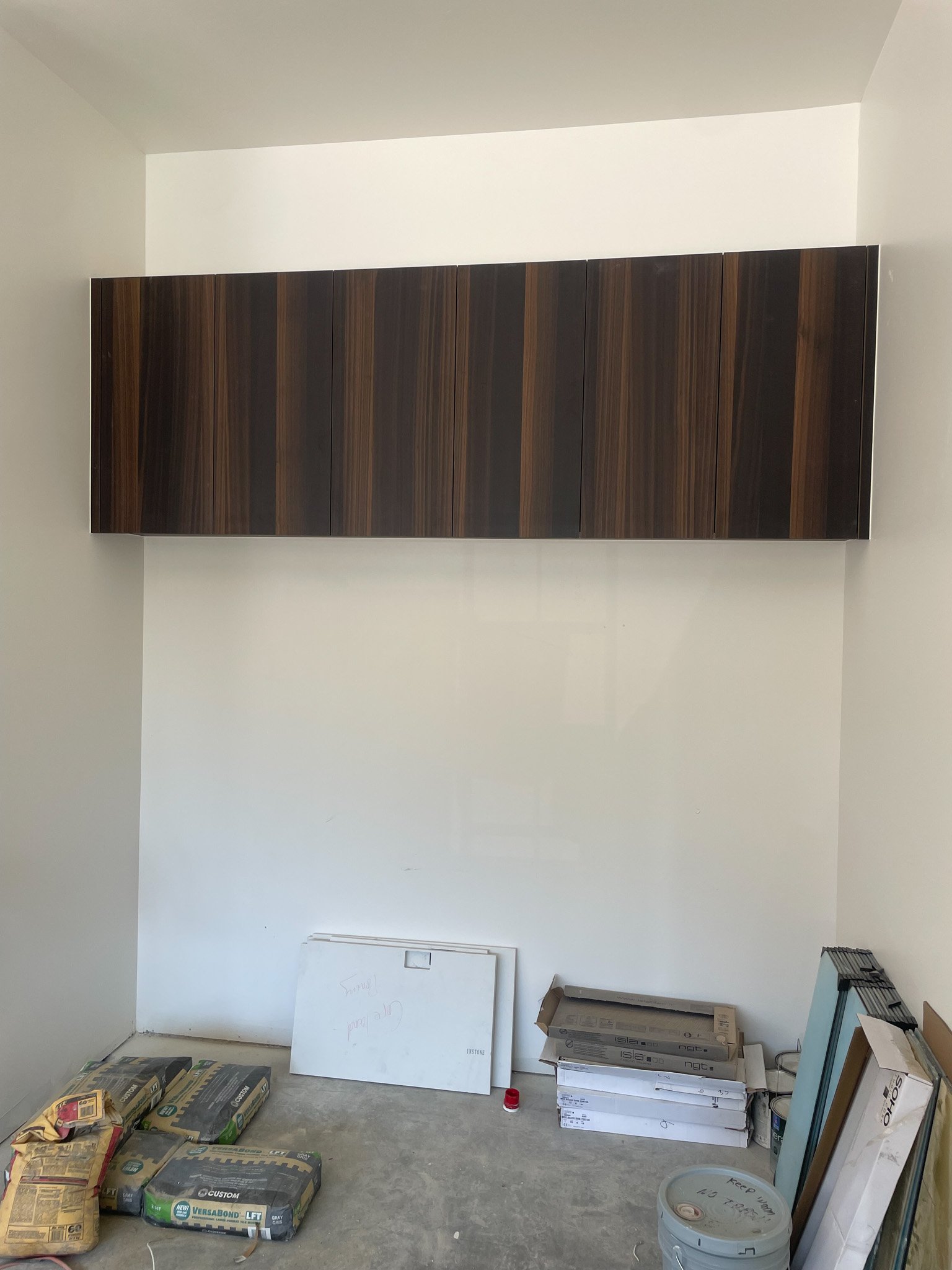 Room with white walls and concrete floor, with a dark wood cabinet mounted on the wall, and various construction materials and boxes on the floor.