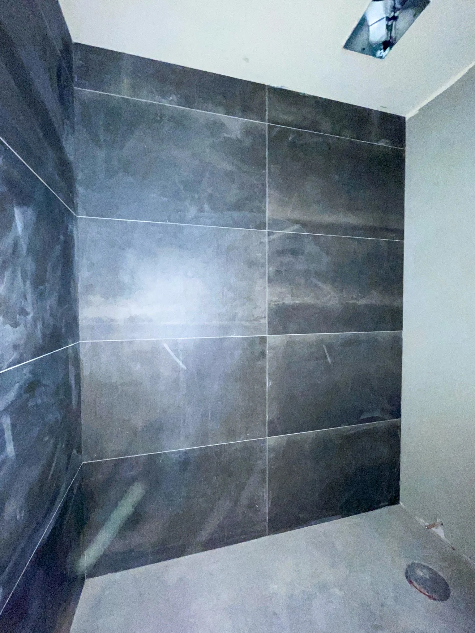 Close-up of a bathroom corner with dark gray tiles on the wall and unfinished concrete on the floor, plumbing hole in the floor, and a partially installed ceiling fixture.