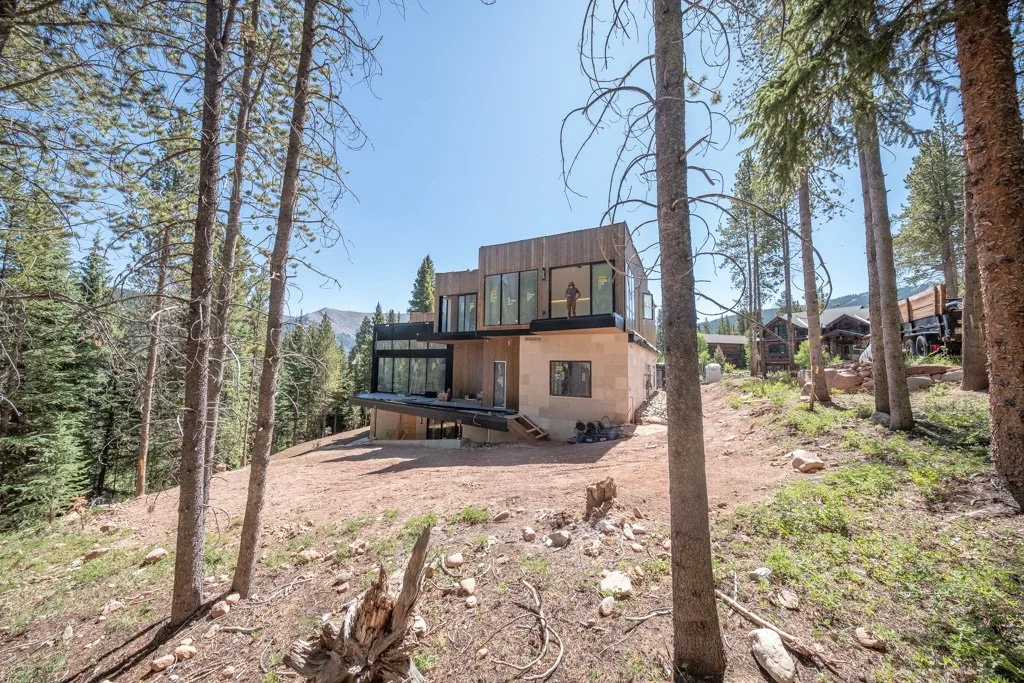 Modern house overlooking a forested hillside with tall pine trees, clear blue sky, and some neighboring houses visible in the distance.