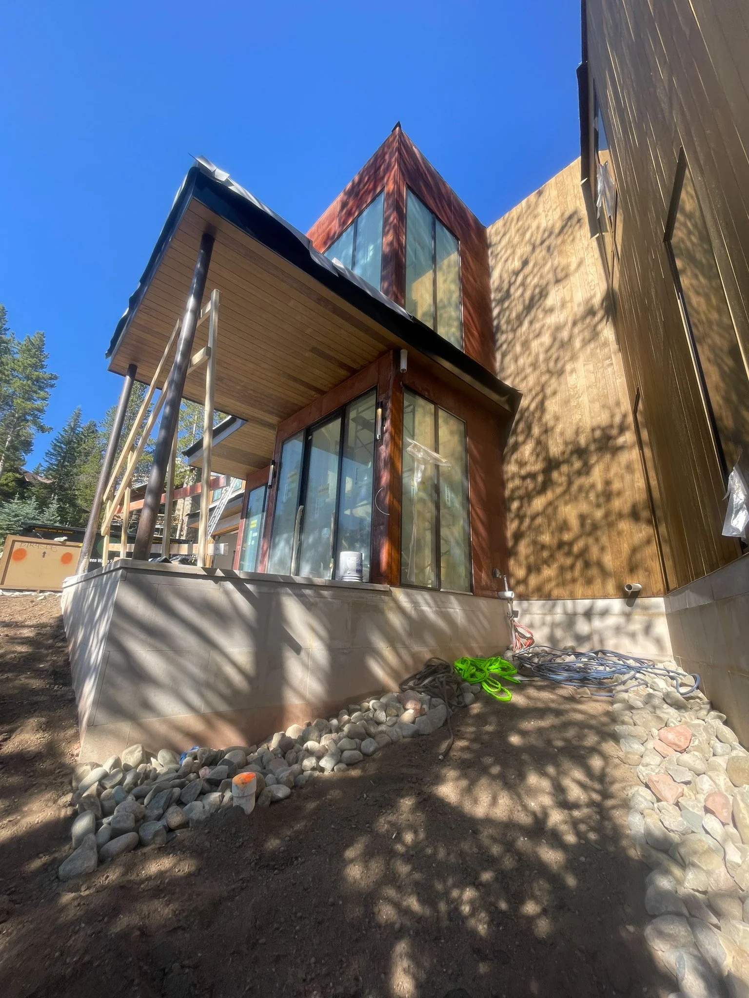 Construction site of modern house with large glass windows and wooden exterior, surrounded by trees, with shadows cast on the ground and construction materials nearby.