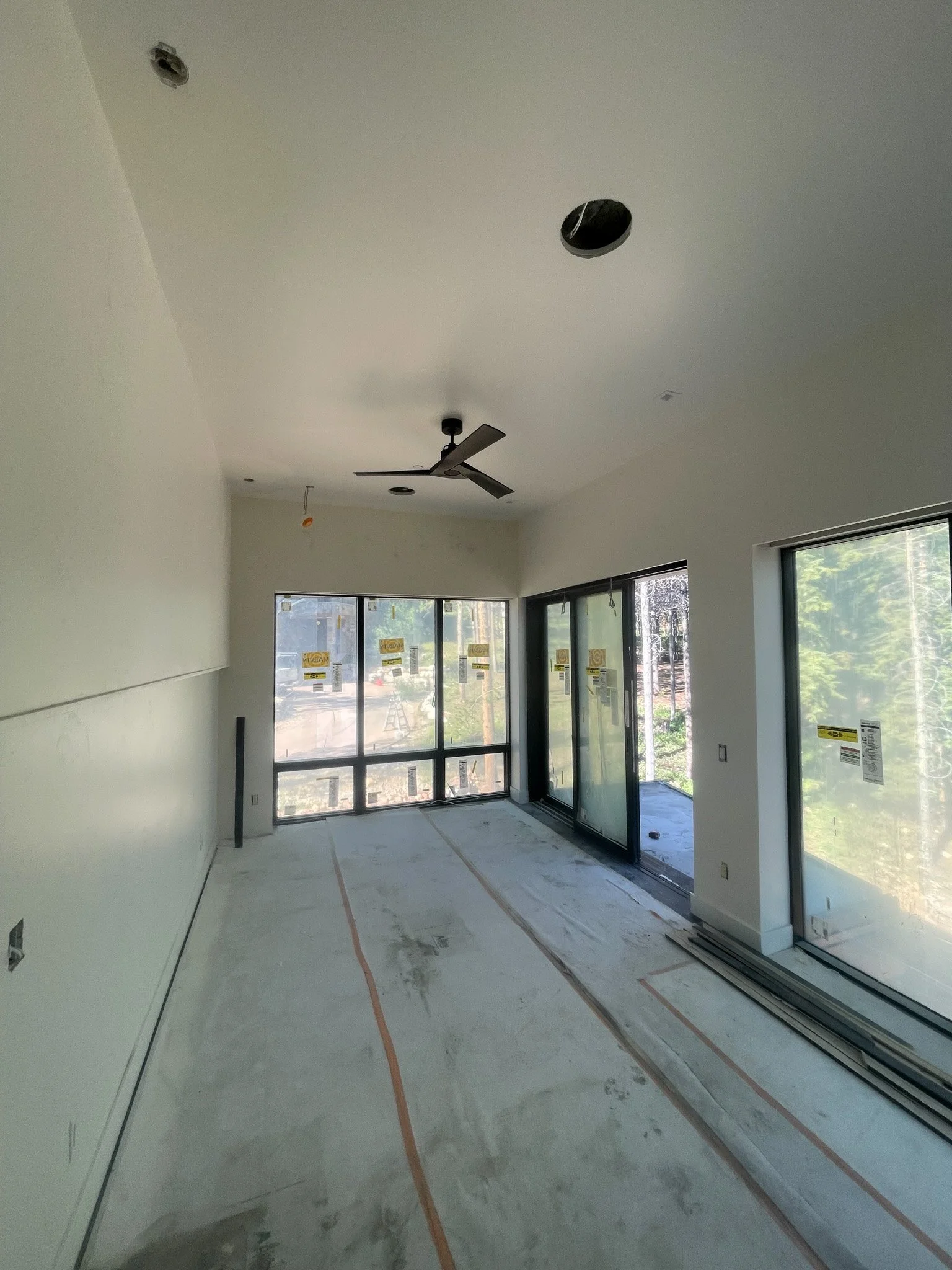 Interior of a house under construction with white walls, large windows, a ceiling fan, and no ceiling lights installed yet.