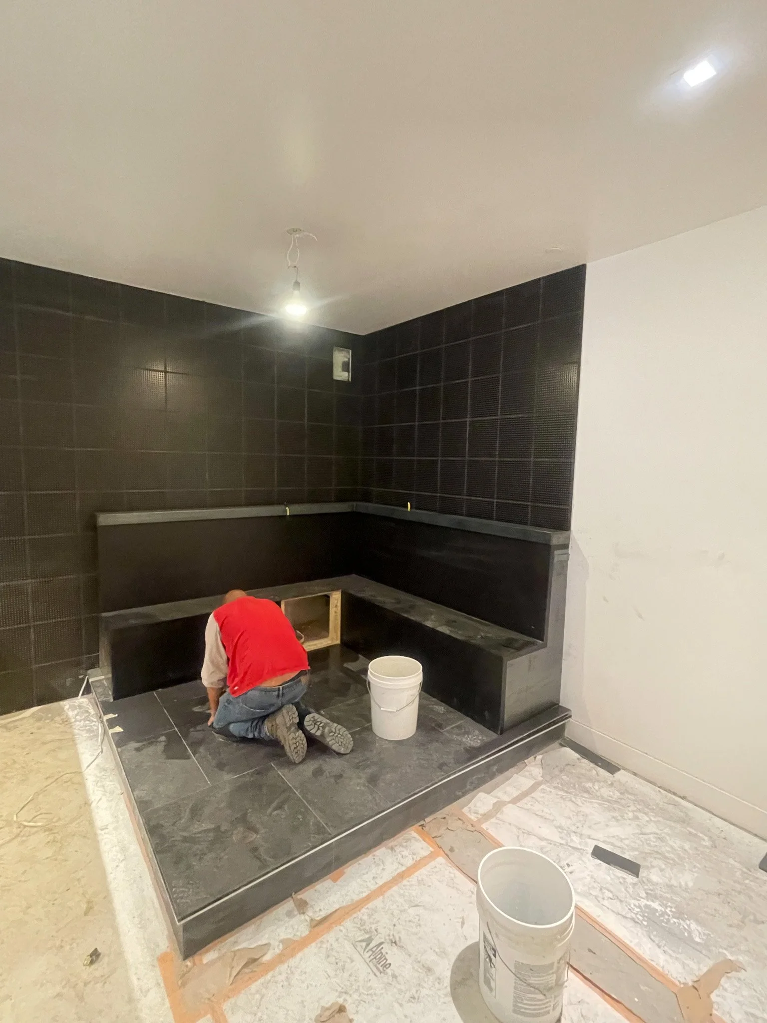 Worker installing black tile flooring in a corner of a room with black tiled walls and a white ceiling, with construction materials and tools around.