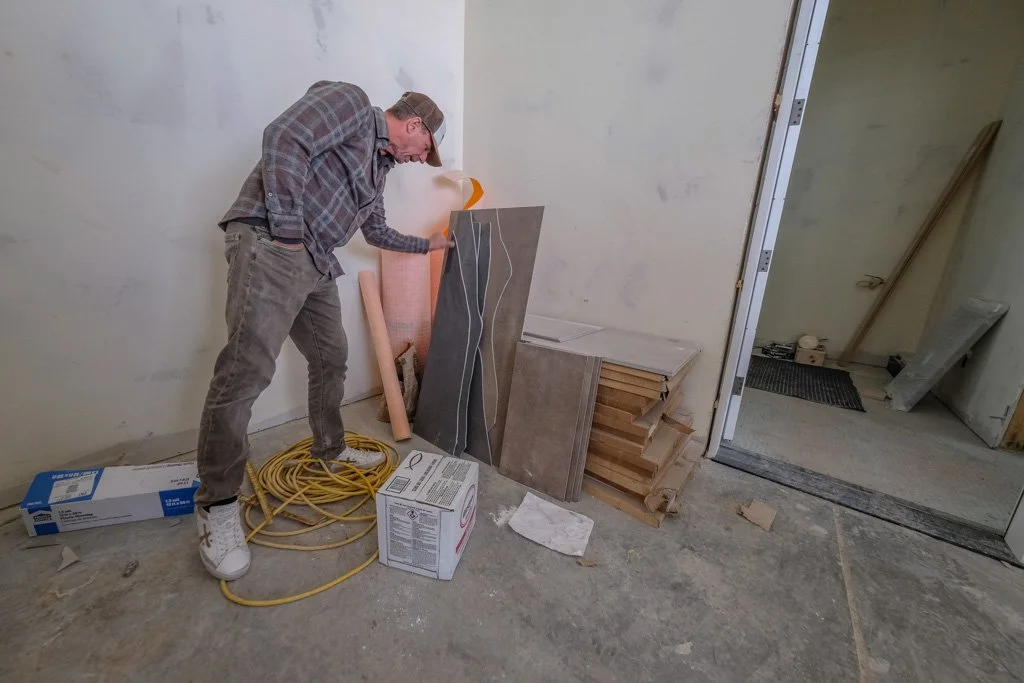A man in a plaid shirt, gray pants, and a cap is working on construction or renovation in a room, handling some wiring or cables with wood shingles, and construction materials and tools around him.
