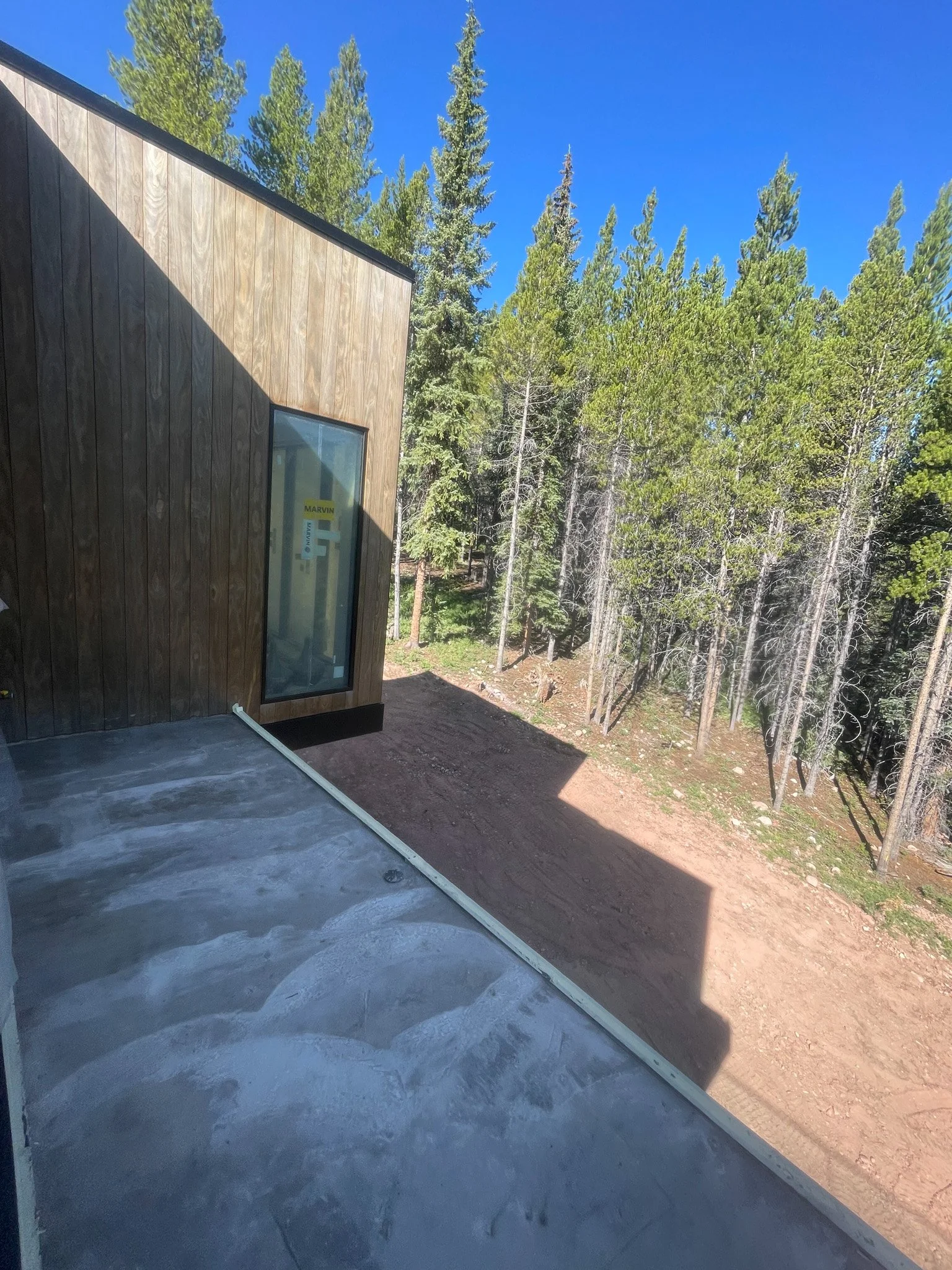 Newly constructed balcony with concrete floor, wooden exterior walls, a glass door, and a forested hillside view with pine trees under a clear blue sky.