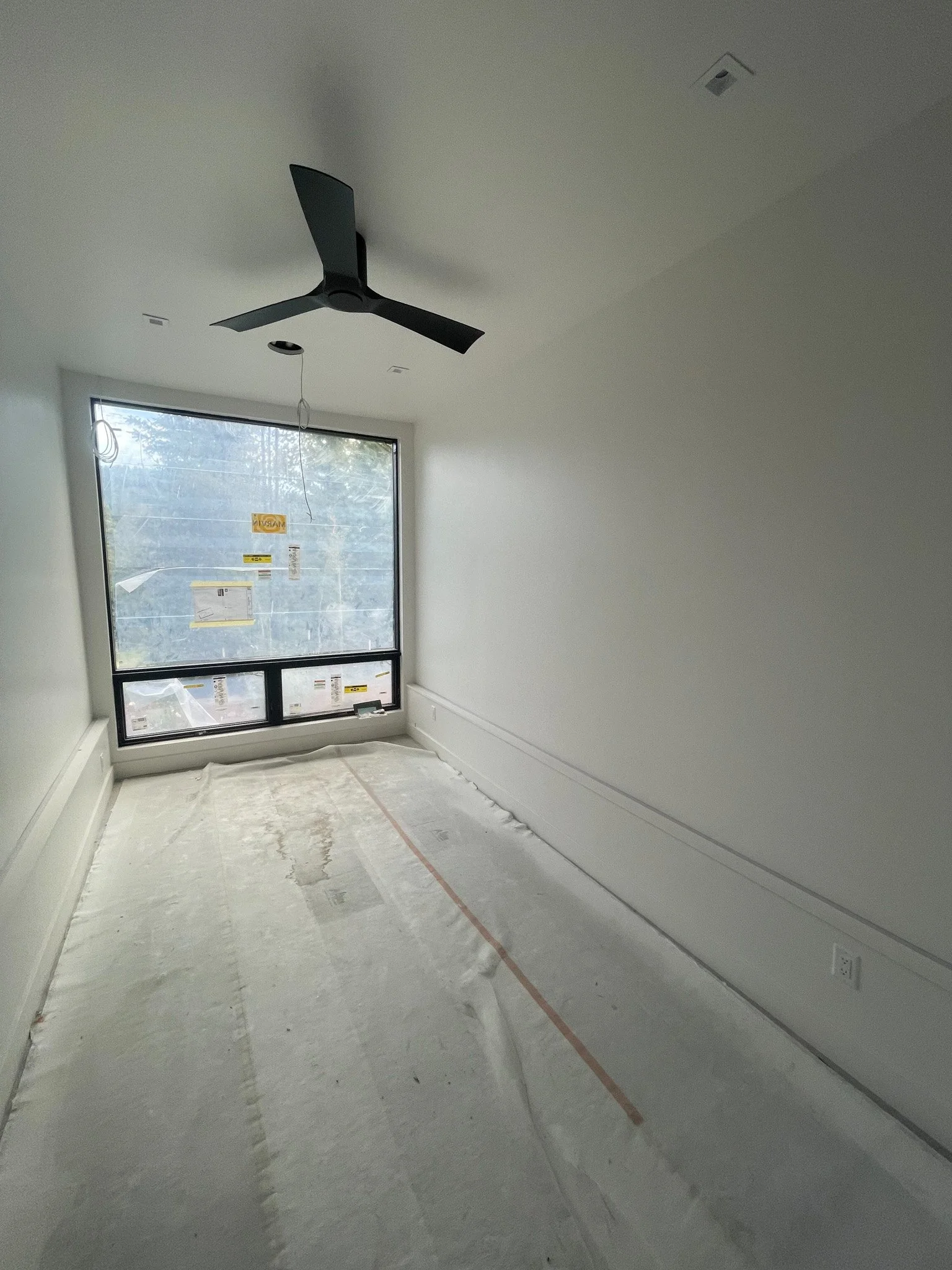 Empty room under construction with a large window, a black ceiling fan, and white walls. The floor is covered with protective plastic.