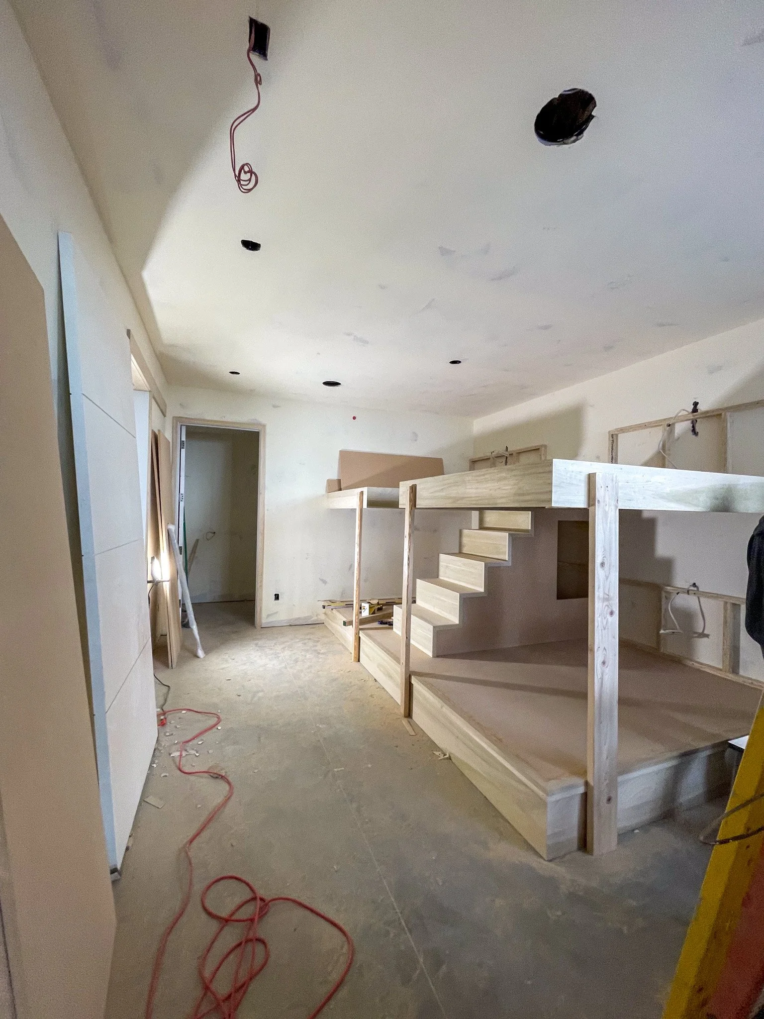Room under construction with unfinished wooden staircase and drywall, ceiling with cutouts and hanging wires.