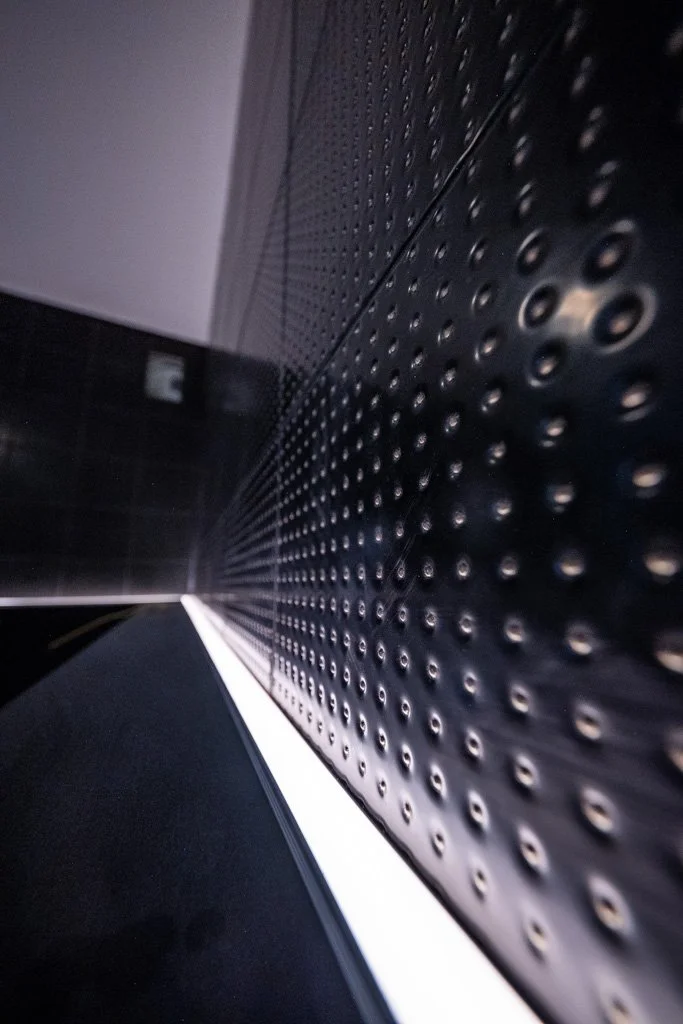 Close-up of a textured black metallic surface with small circular indentations, angled toward a wall and a light source in the background.