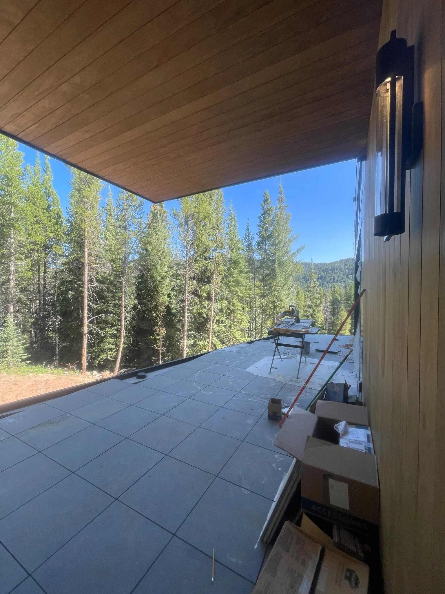Balcony under construction with tiled floor, surrounded by a forest of tall green trees, and a clear blue sky visible in the background.
